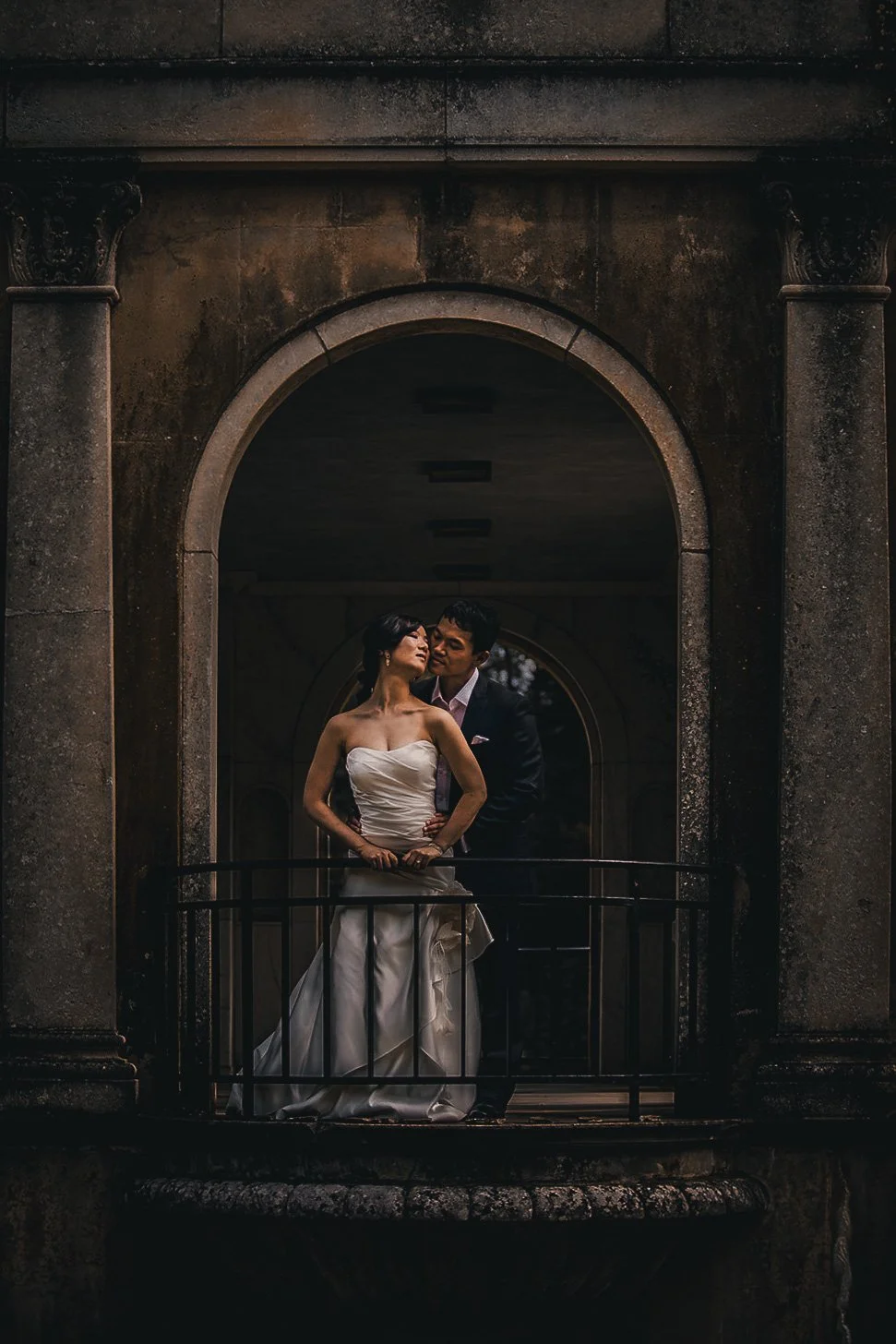A bride and groom standing under an arched stone structure, with the bride in a strapless white wedding gown and the groom in a black suit, sharing an intimate moment.