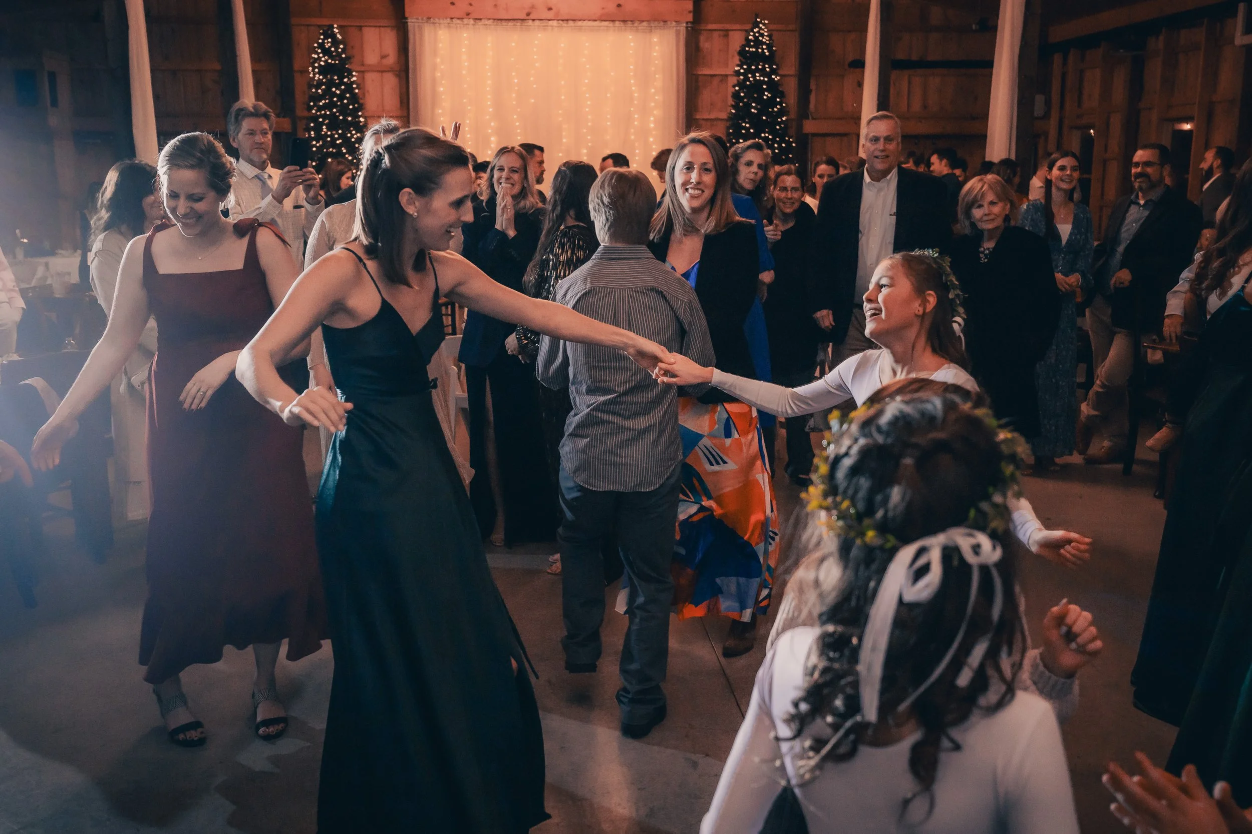 People dancing and celebrating at a festive event in a decorated wooden venue with Christmas trees and string lights.