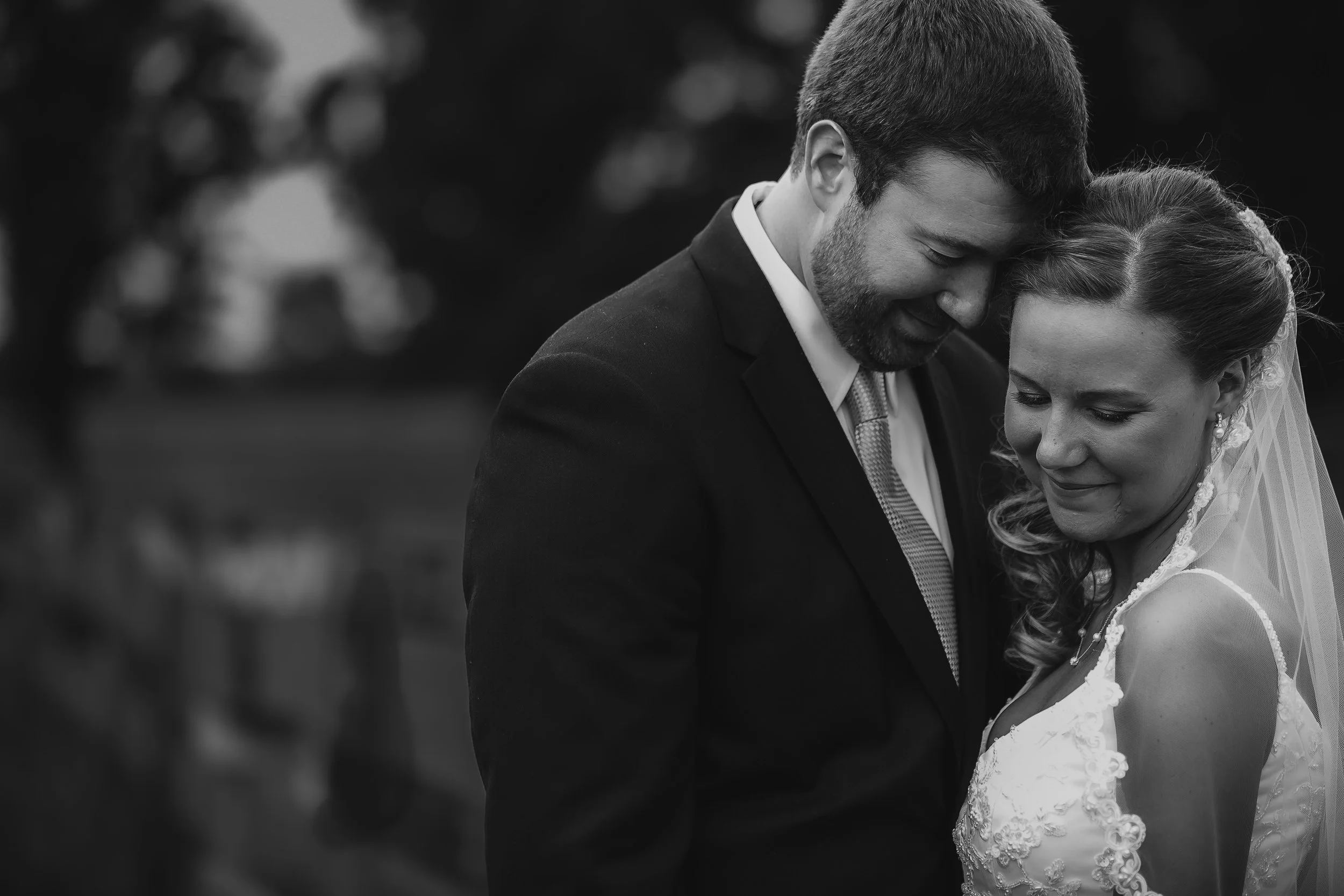 A black and white photograph of a man in a suit and a woman in a wedding dress, with their foreheads touching and eyes closed, sharing an intimate moment outdoors.