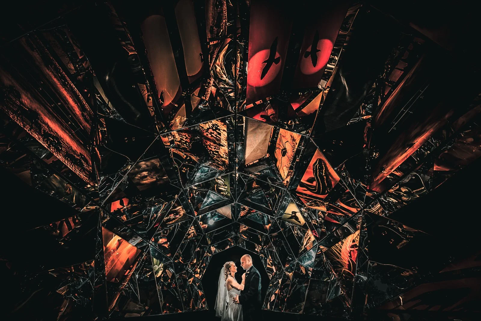 A bride and groom standing close together inside a dark, reflective, and geometrically intricate room, gazing at each other, with colorful distorted reflections surrounding them.
