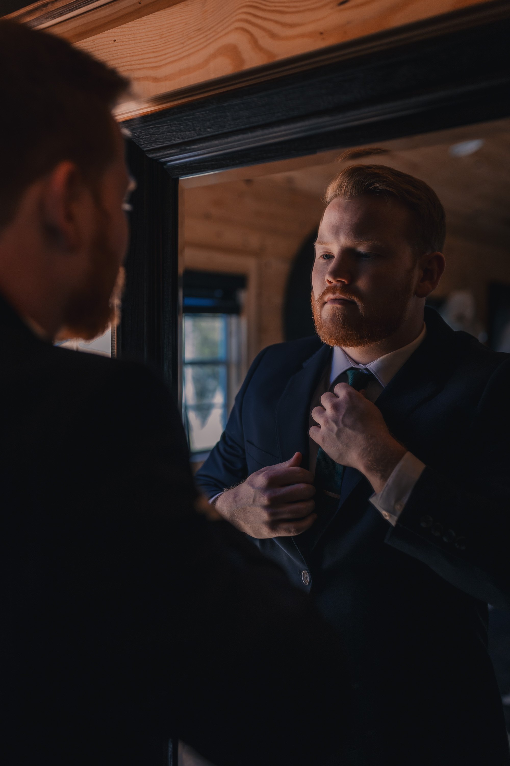 A young man with a beard adjusting his tie in front of a mirror, wearing a dark suit, in a wooden interior room with natural light from a window.