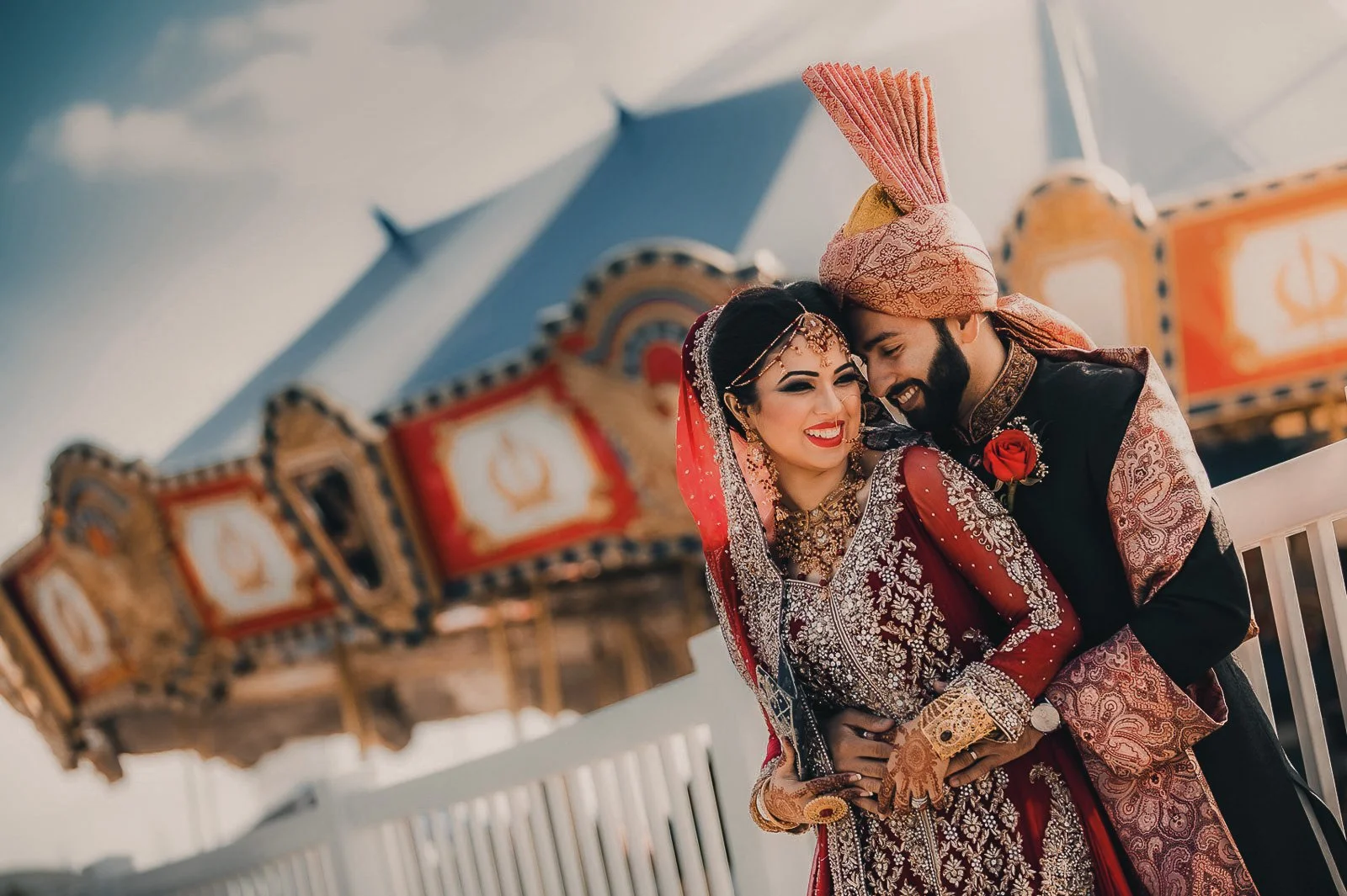 A South Asian couple in traditional wedding attire at an amusement park, both smiling and embracing.