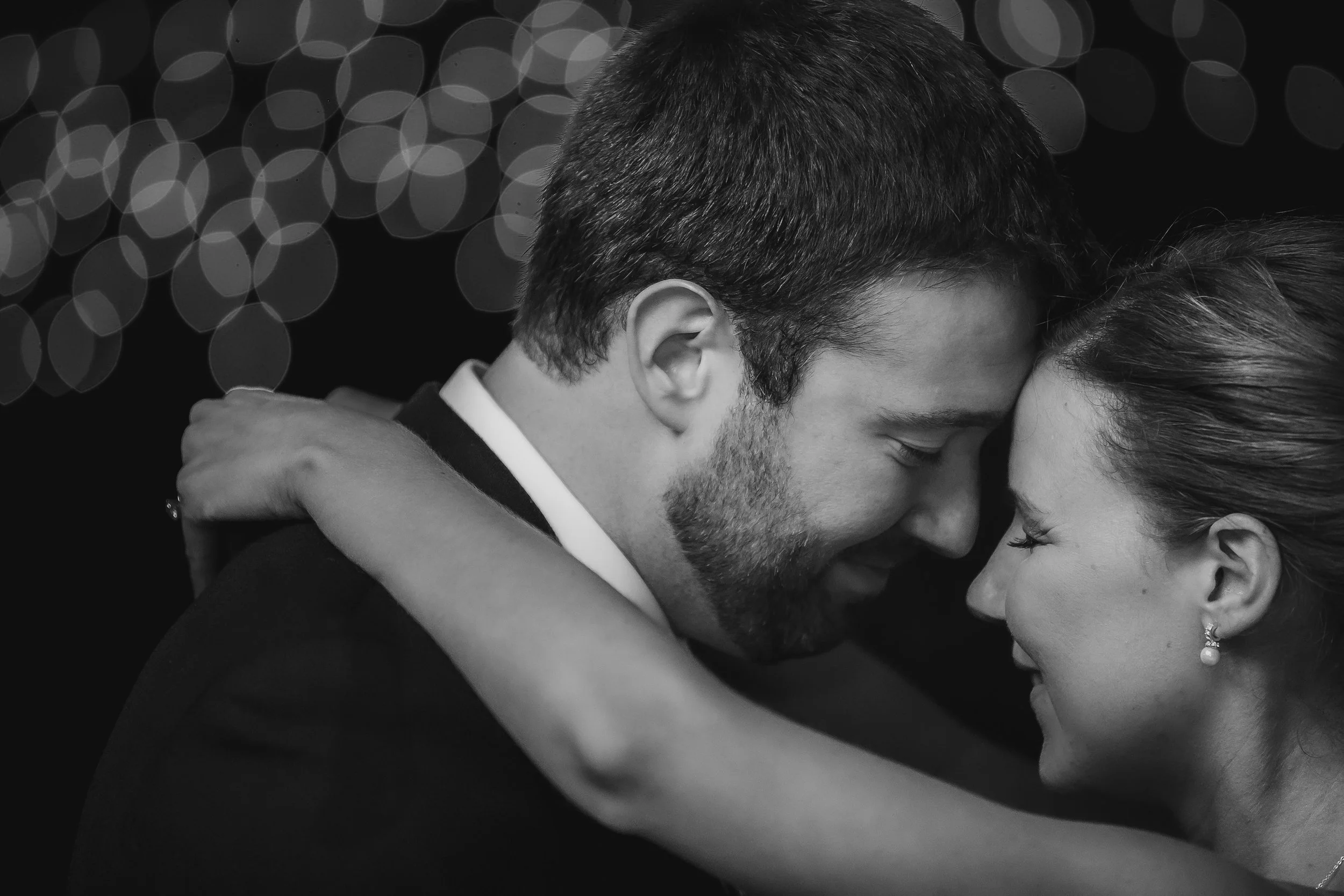 A black and white photo of a couple sharing an intimate moment, their faces close together, with the man's arms around the woman's shoulders.