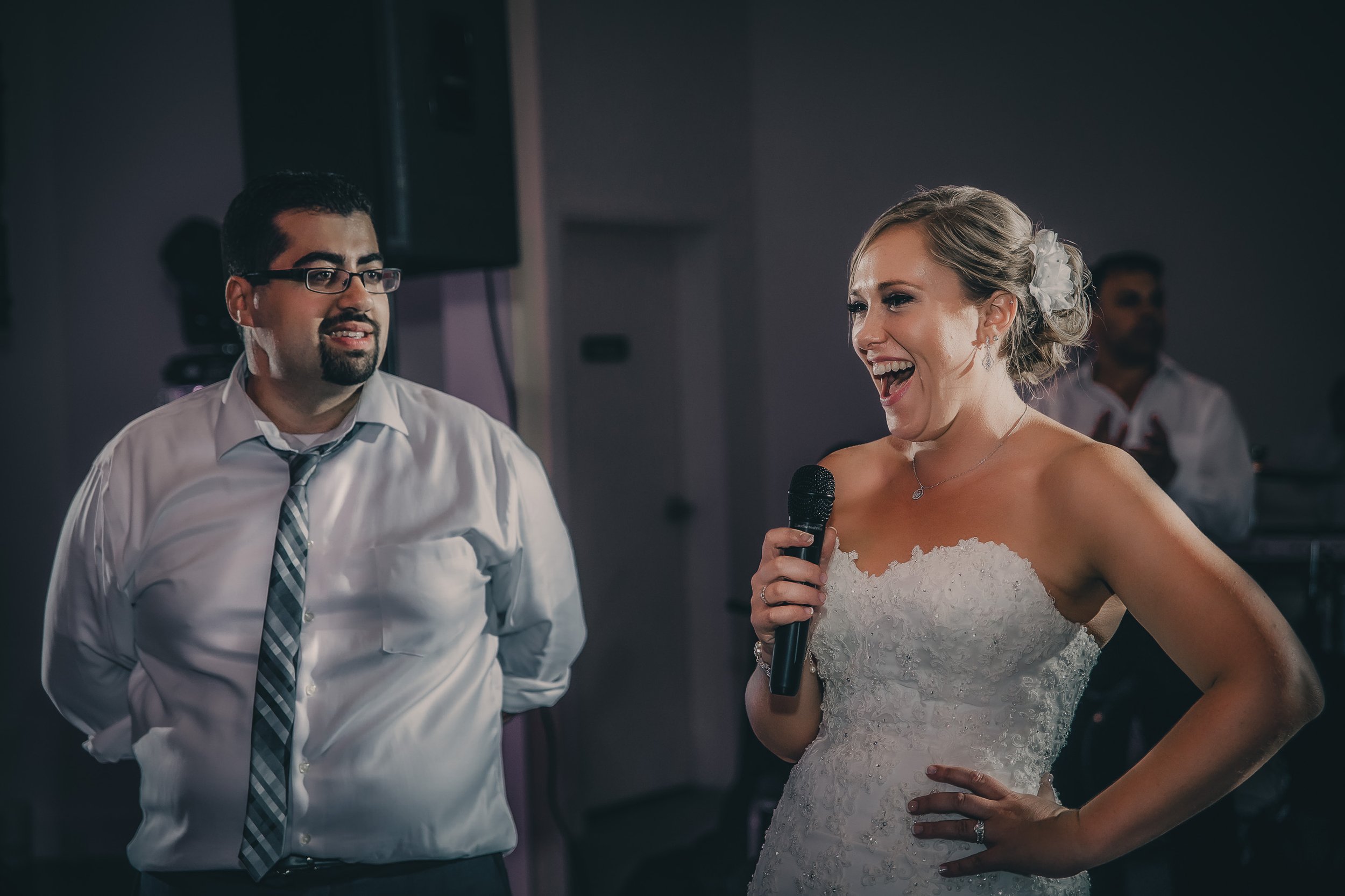 A bride in a white wedding dress holding a microphone and smiling while talking, standing next to a man in a white shirt and striped tie at a wedding reception.