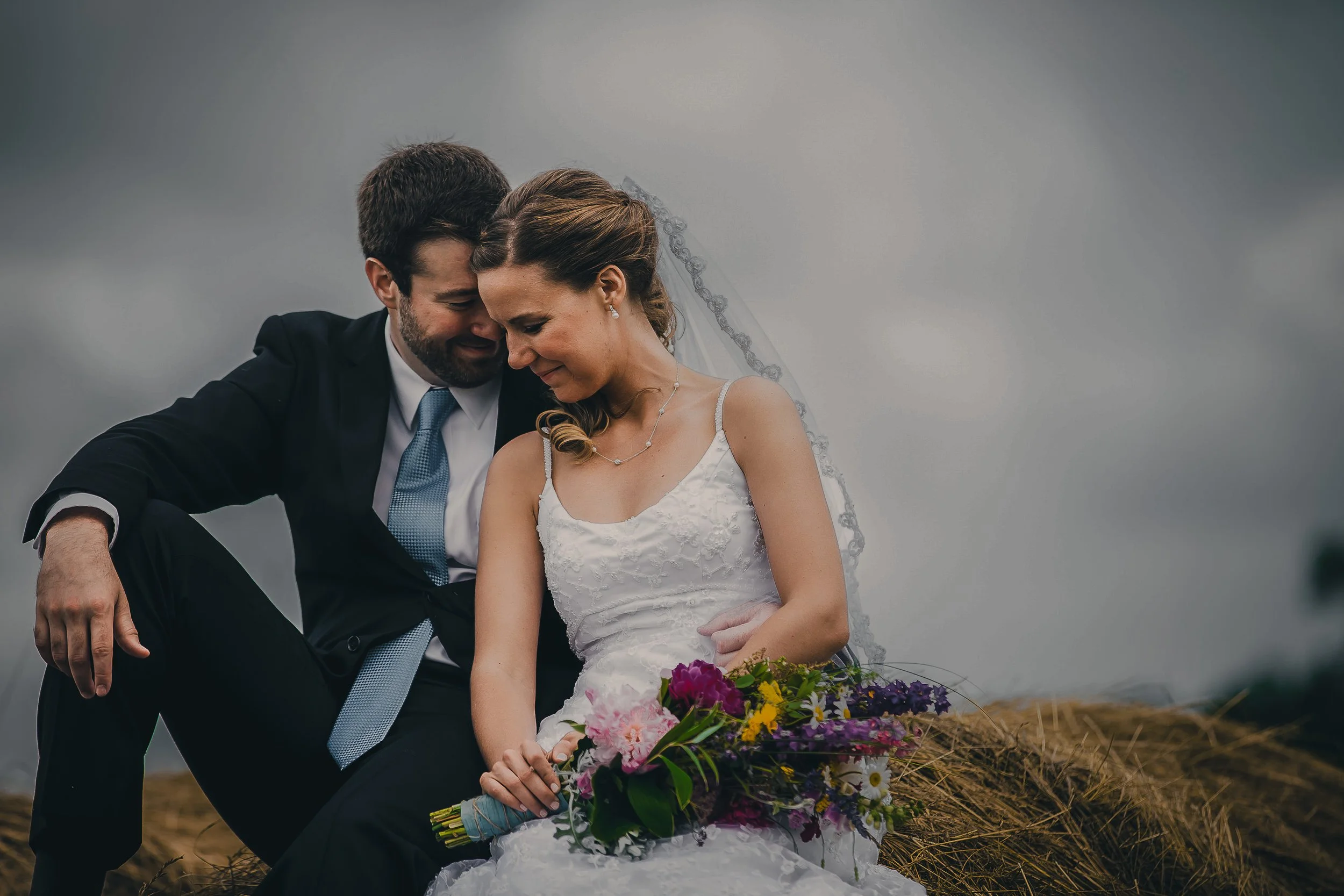 A wedding couple sitting close together outdoors, with the groom in a black suit and the bride in a white wedding dress holding a colorful bouquet of flowers, both with closed eyes and gentle smiles.