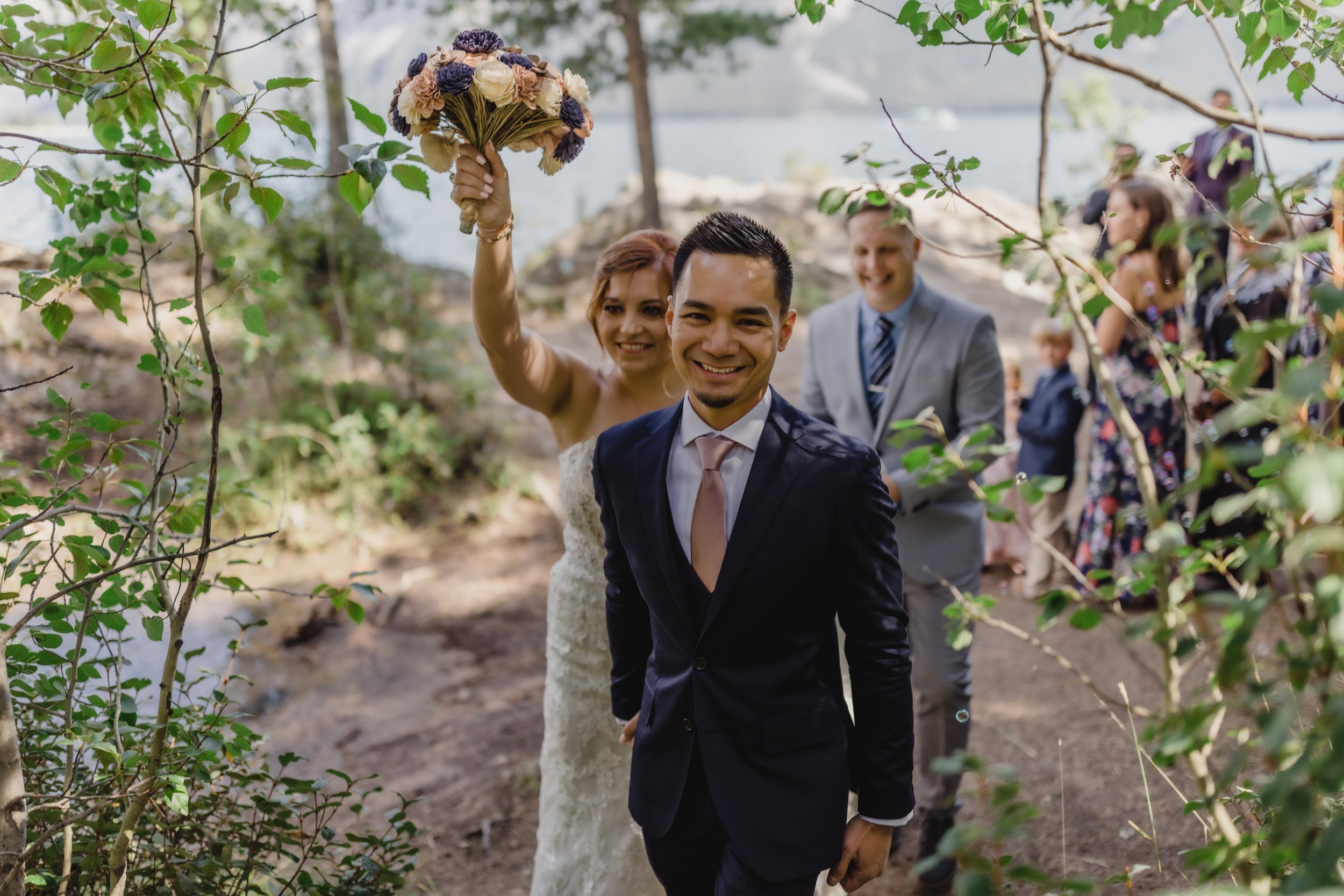 A group of people, including a bride in a wedding dress holding a bouquet up, and a groom in a suit, are walking outdoors through trees by a body of water, celebrating a wedding.