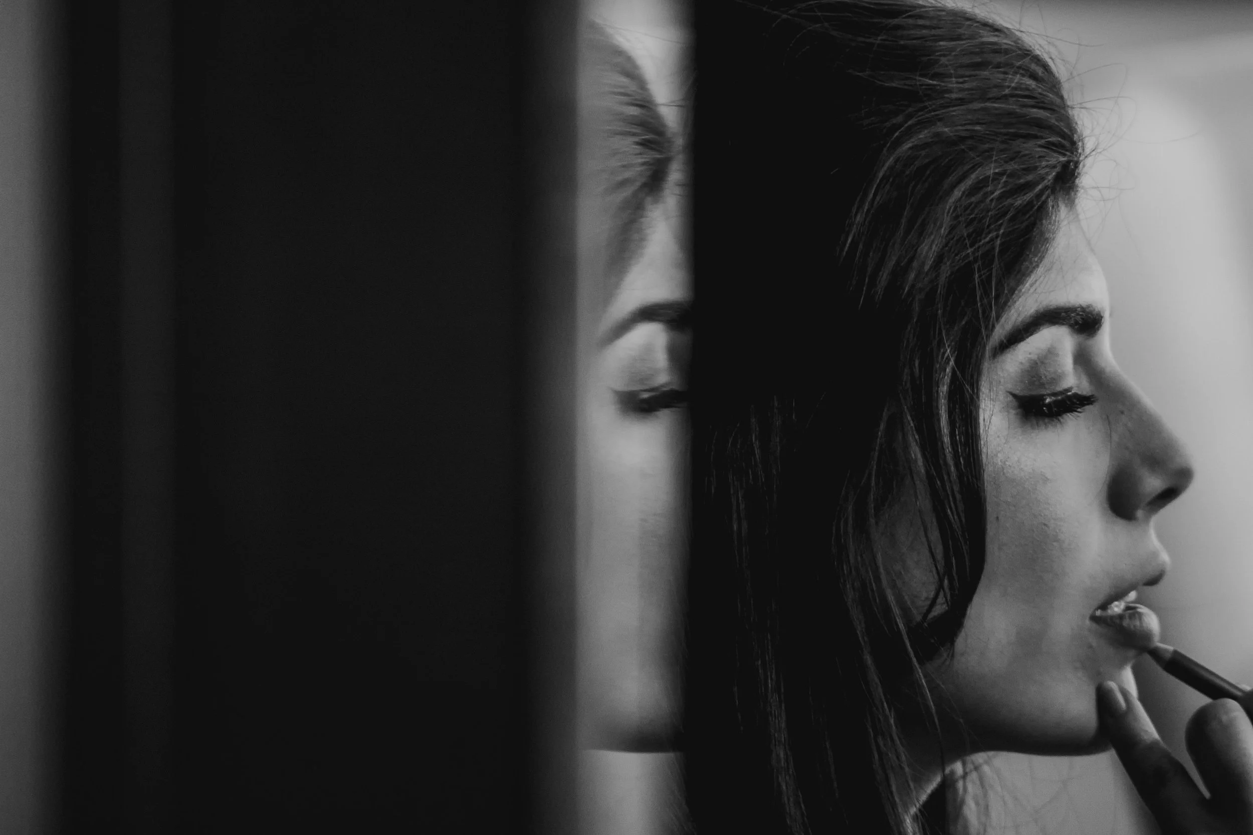A black and white photo of a woman with dark hair, applying lipstick with her eyes closed.