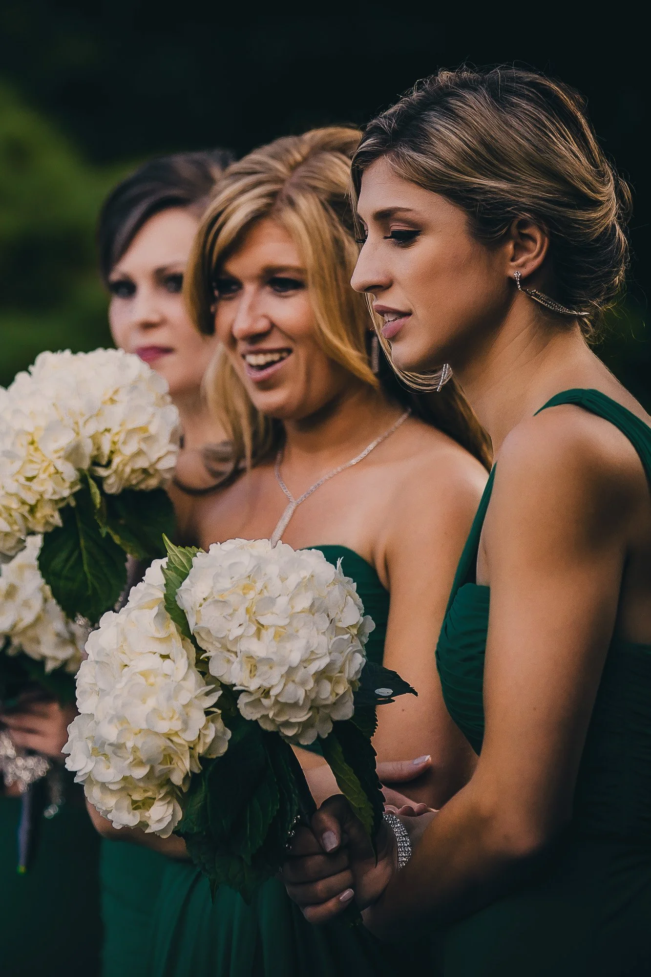 Three women in green dresses holding white hydrangea bouquets during an outdoor event.