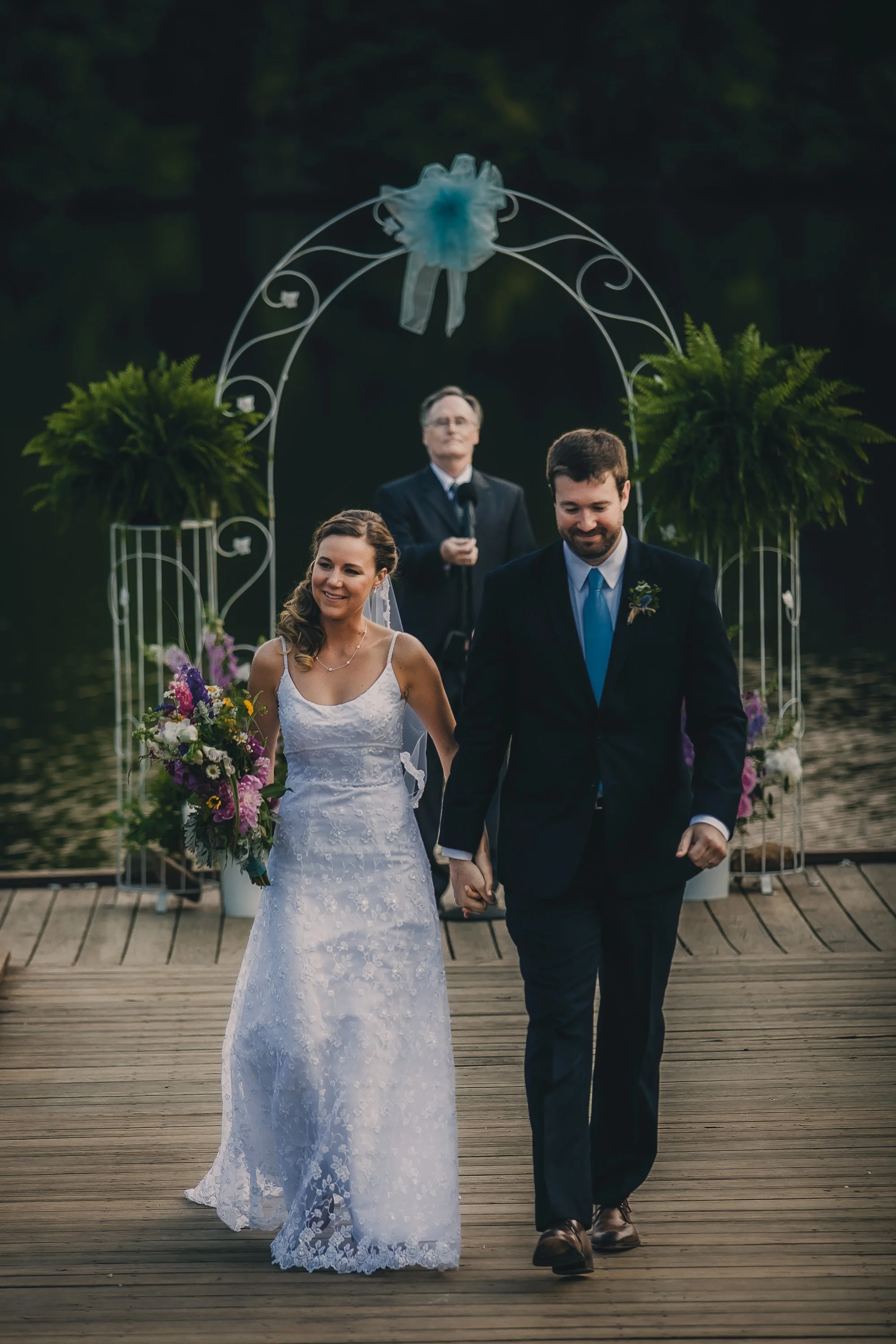 A newlywed couple walking hand in hand on a wooden dock during their wedding ceremony, with an officiant in the background and decorated with flowers and greenery.