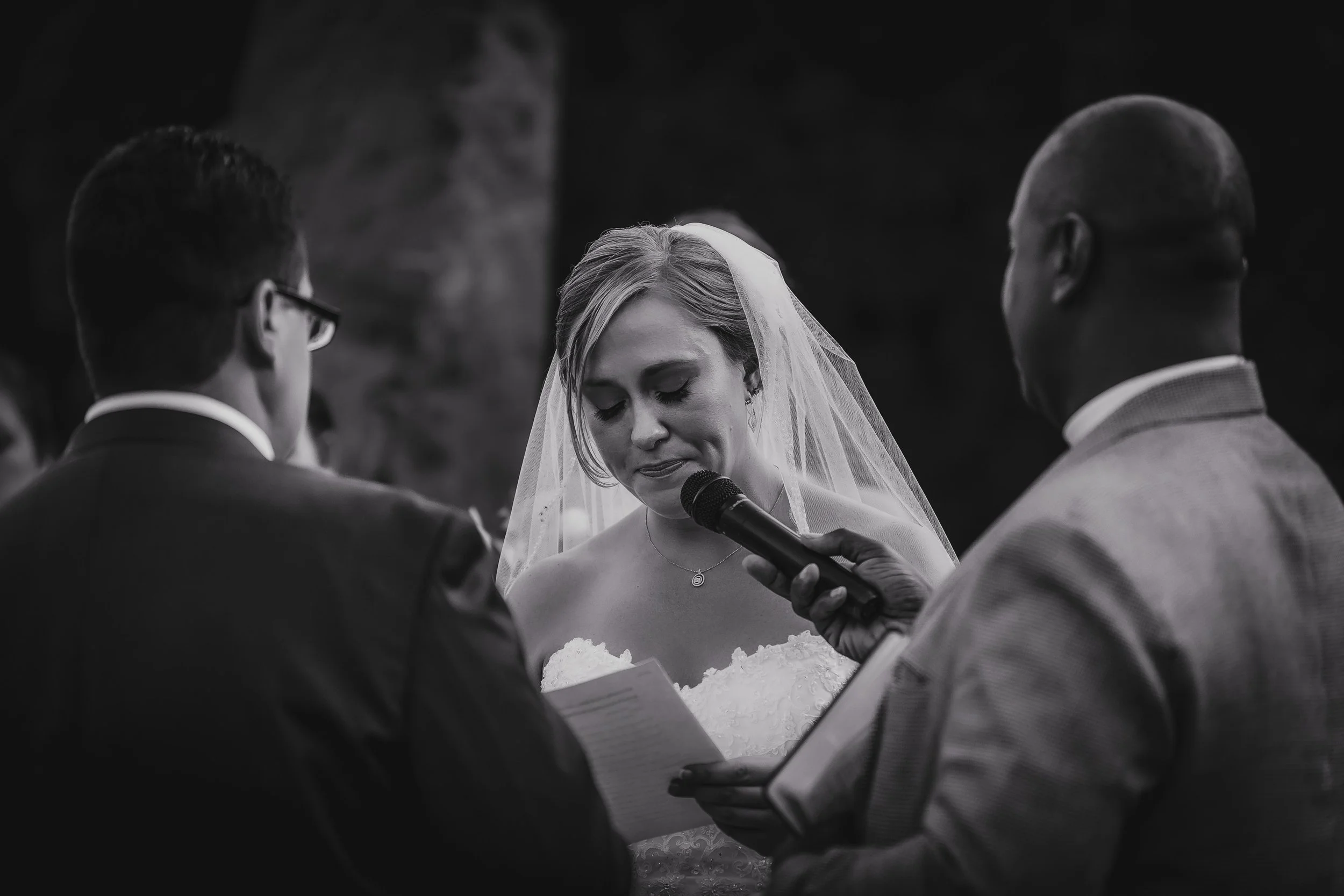 A black-and-white photo of a wedding ceremony with a bride in a wedding dress and veil, holding a prayer book, standing between two men. One man is holding a microphone near her face and reading from a paper.