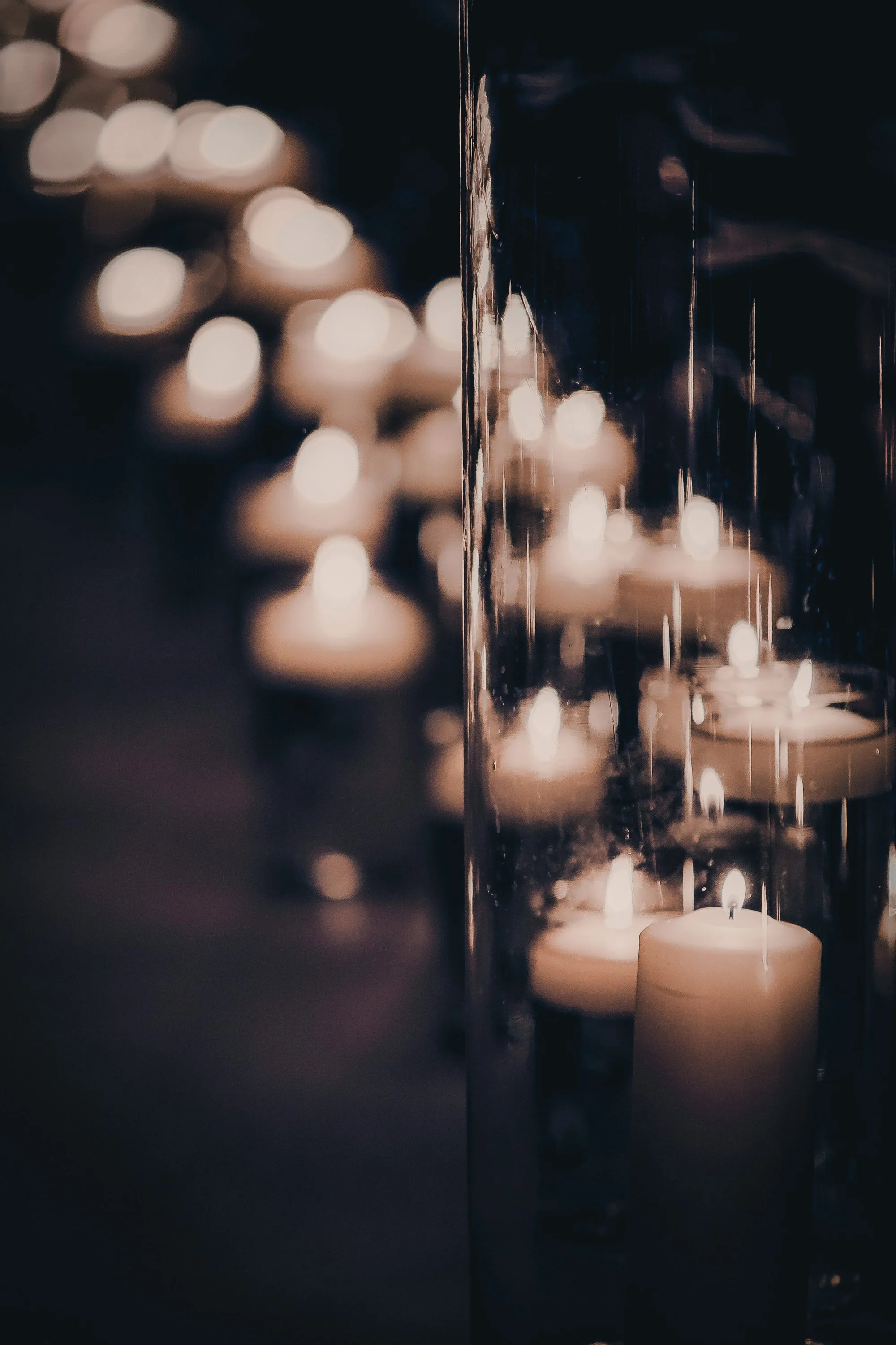 A close-up of a glass candle holder with multiple lit candles inside, with blurred candle lights in the background.