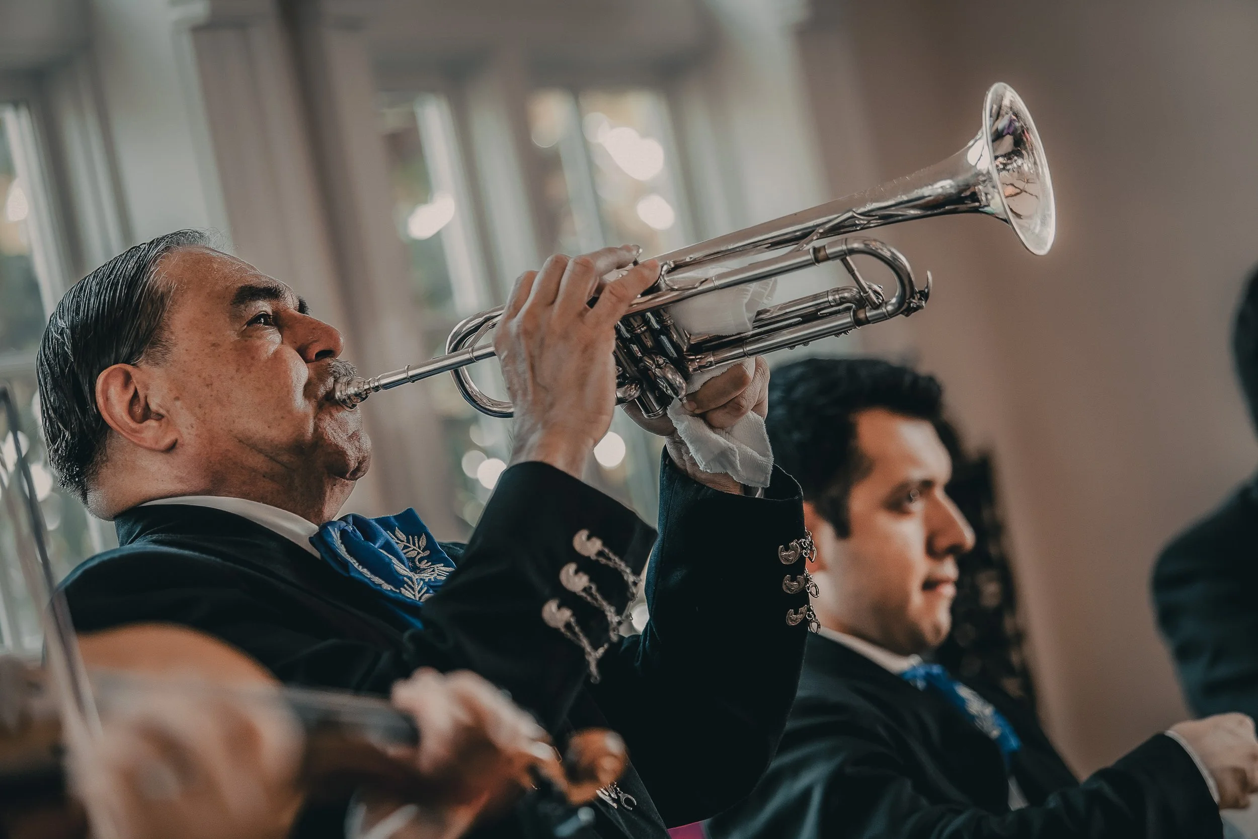 An elderly man playing the trumpet during a musical performance, with two other musicians visible in the background.