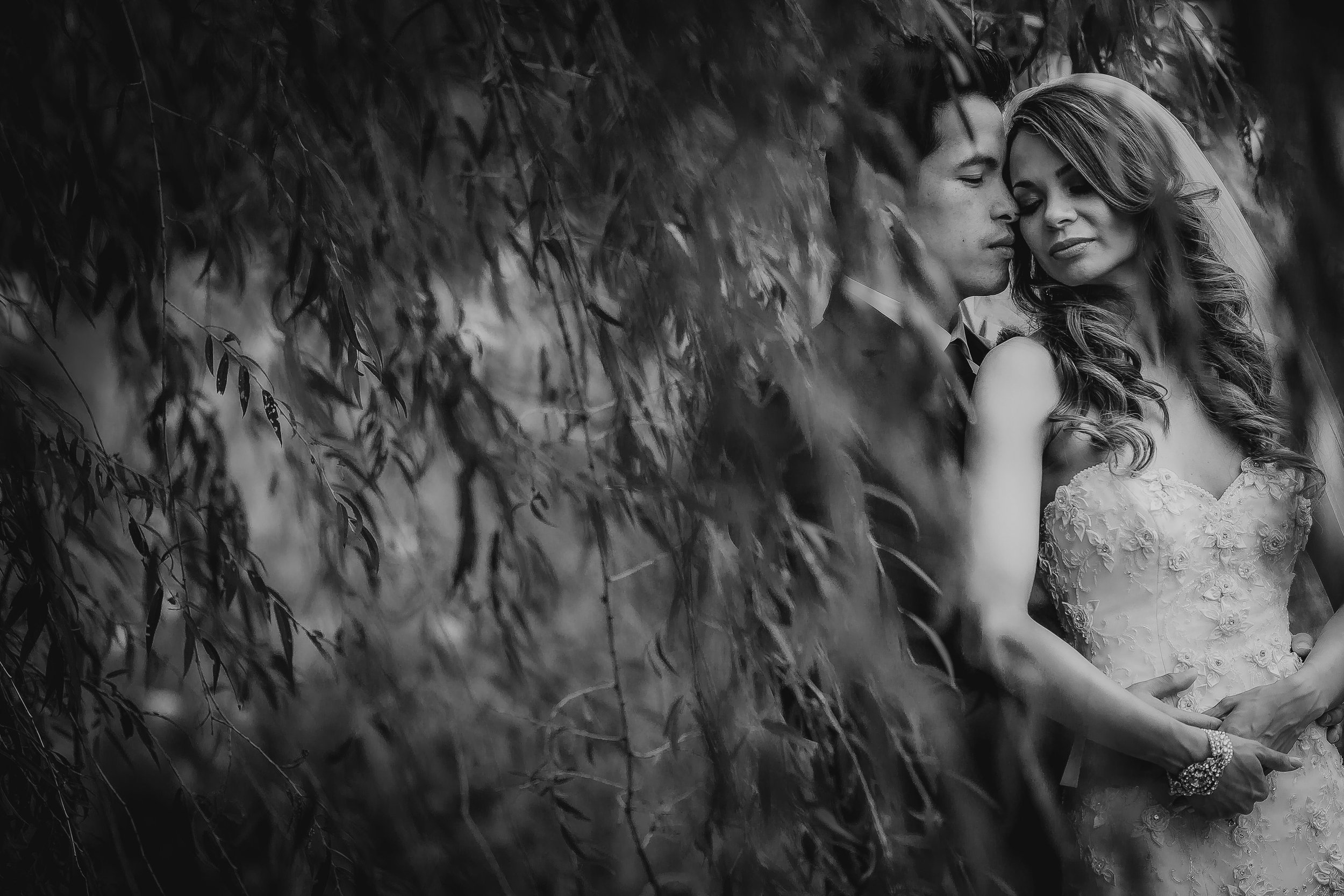 A black and white photograph of a bride and groom standing close together behind a veil of leaves, with the bride wearing a strapless lace wedding dress and the groom in a suit, both with their eyes closed.