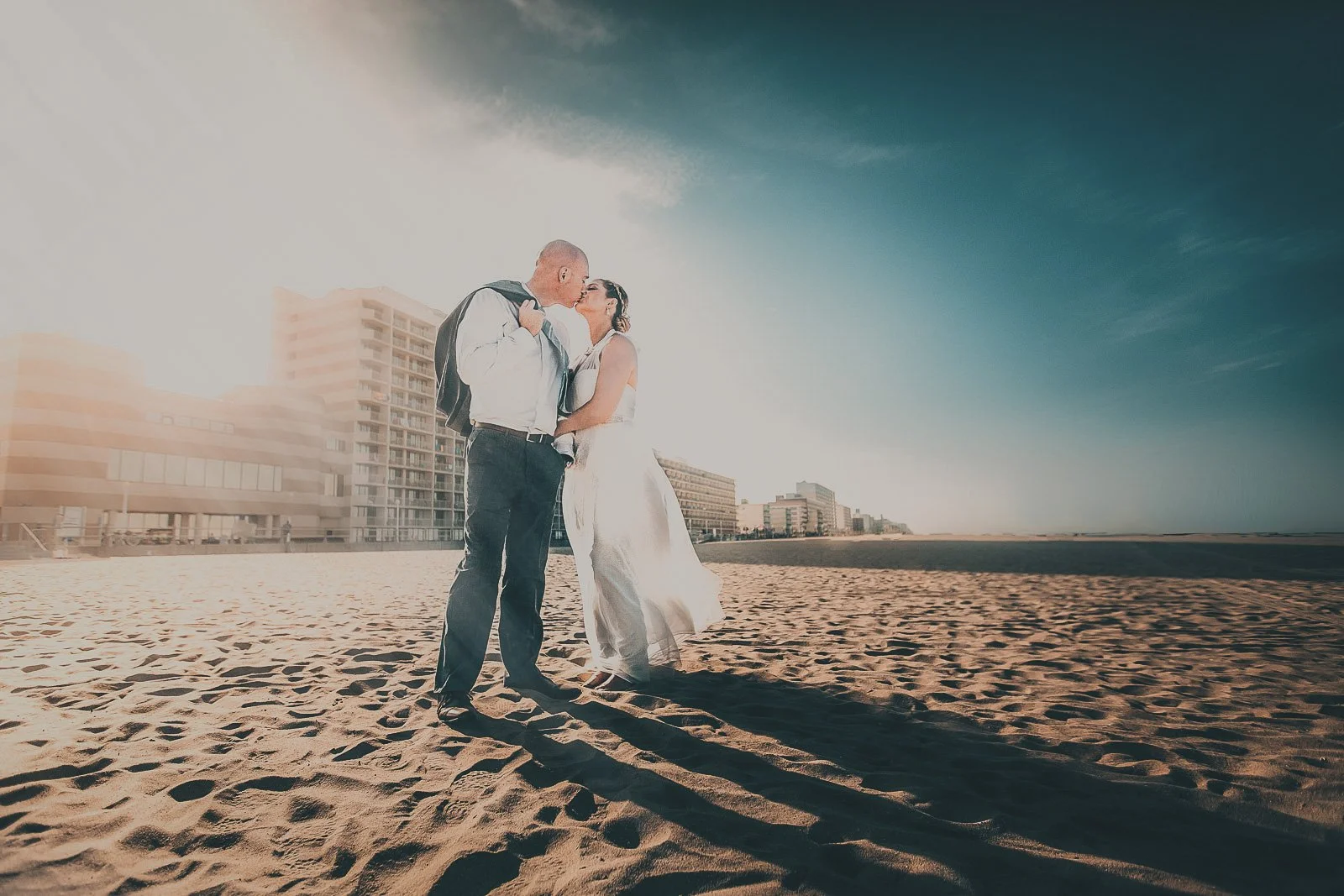 A couple dressed in wedding attire shares a kiss on a sandy beach with city buildings in the background during sunset.