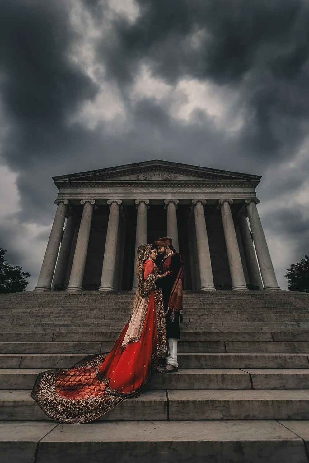 A couple dressed in traditional South Asian wedding attire stands on steps in front of a large classical building with columns, under a cloudy sky.