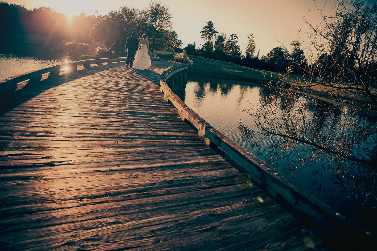 A couple, I believe they are newlyweds, walking on a wooden bridge at sunset over a body of water, with trees in the background.