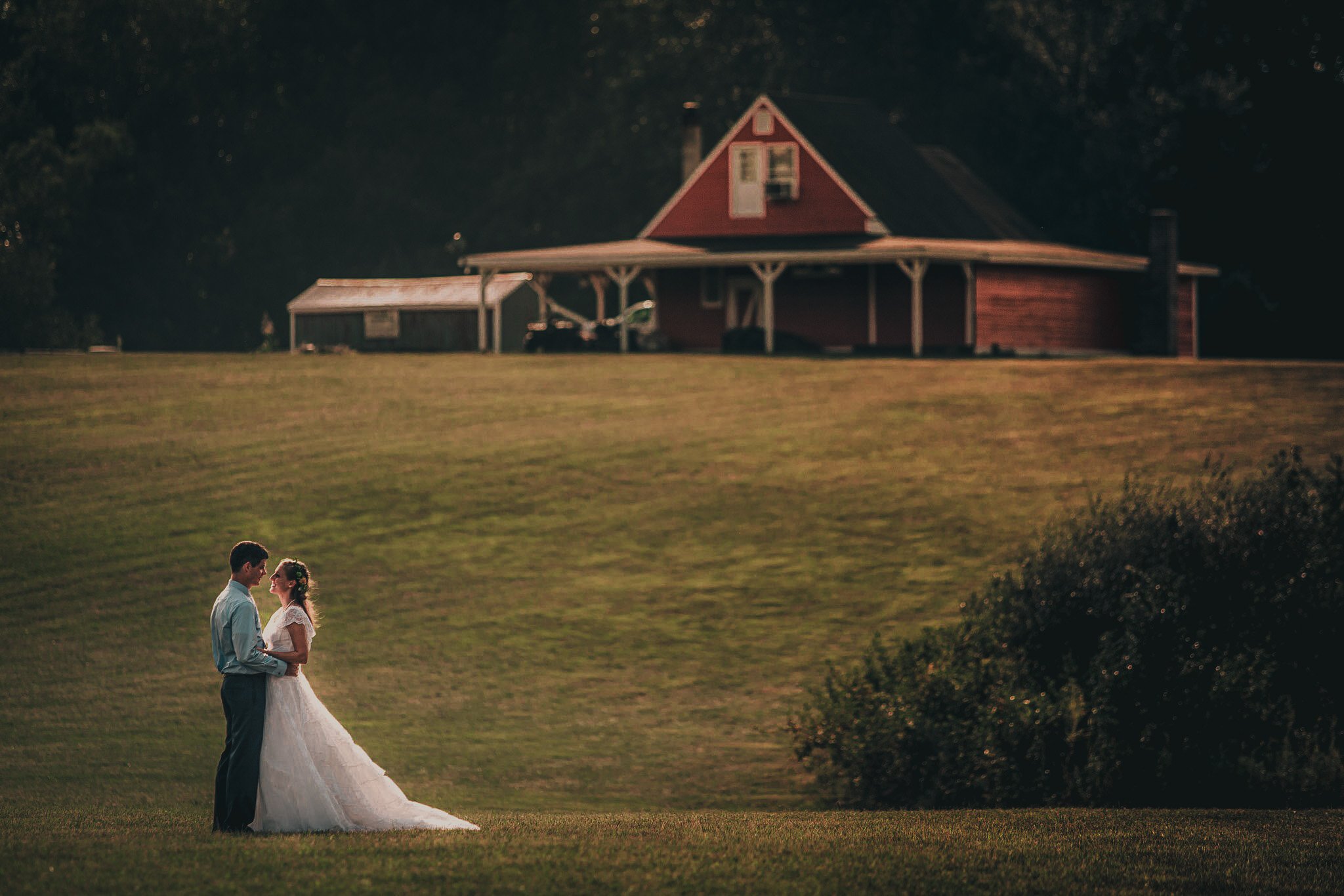 A bride and groom standing close on a grassy field at sunset, with a large red barn and trees in the background.