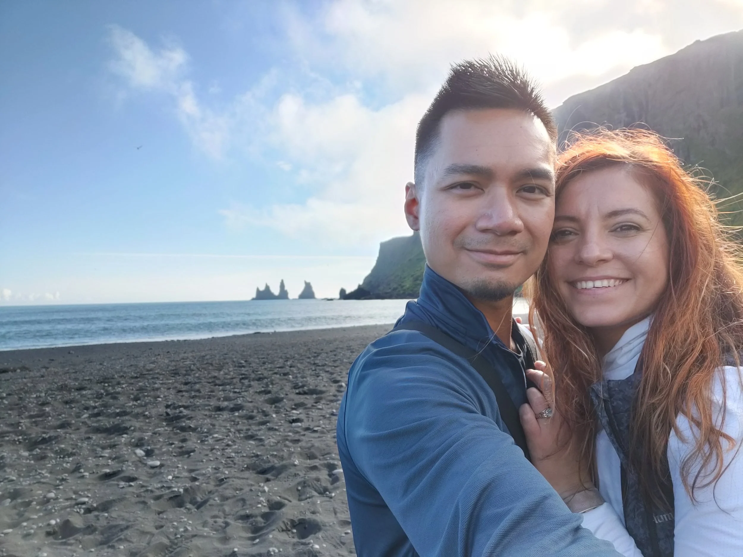 A smiling couple taking a selfie on a black sand beach with mountainous landscape and sea stacks in the background during daylight.