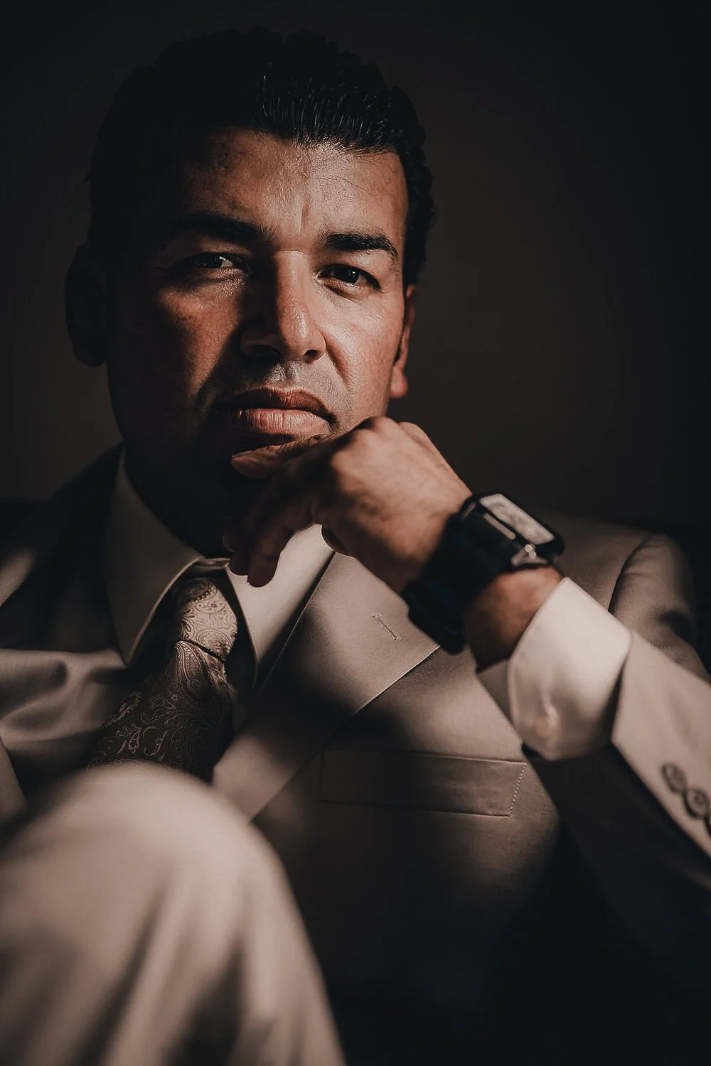 Man in a beige suit with a printed tie, wearing a smartwatch, looking confidently at the camera with his hand near his chin, dark background.