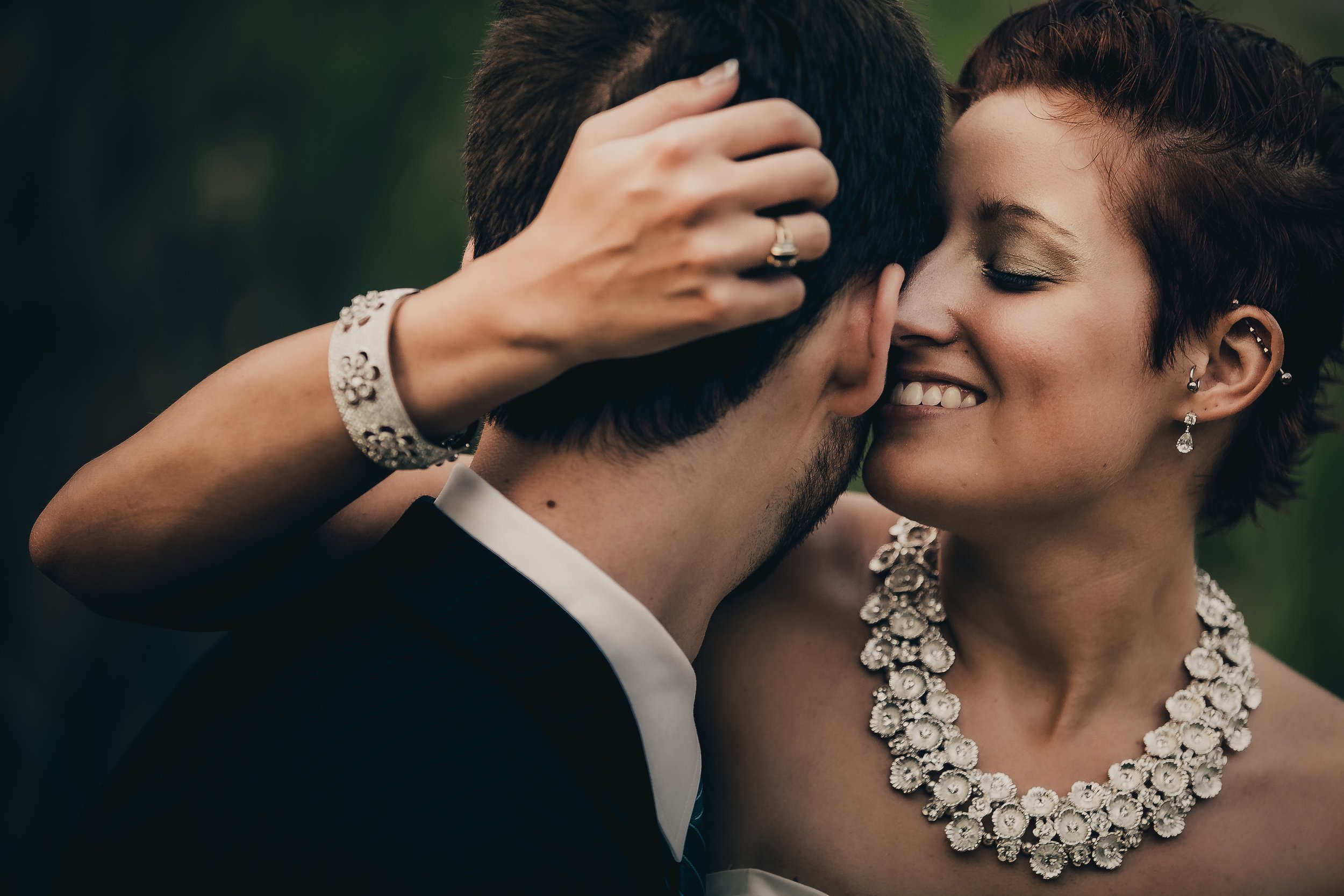 A couple sharing a close, intimate moment, smiling and touching foreheads, dressed in formal attire with jewelry, in an outdoor setting.