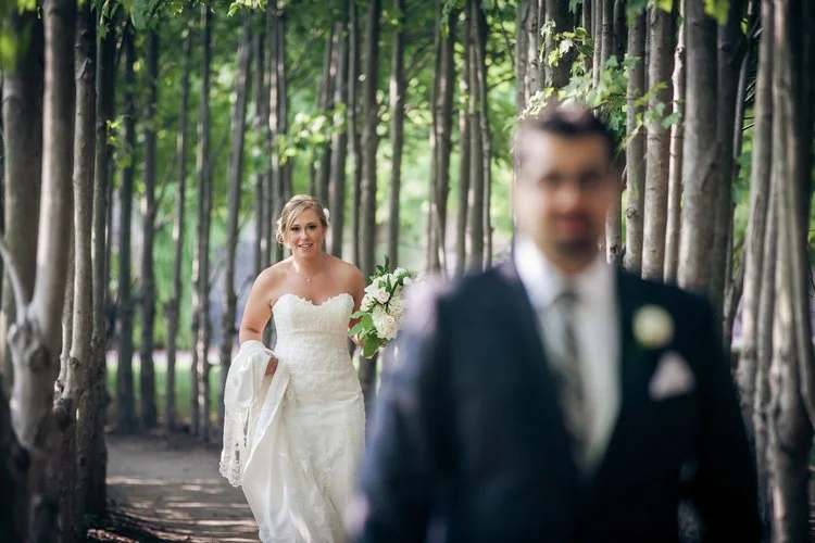 A bride walks up behind the groom during their first look surrounded by trees.