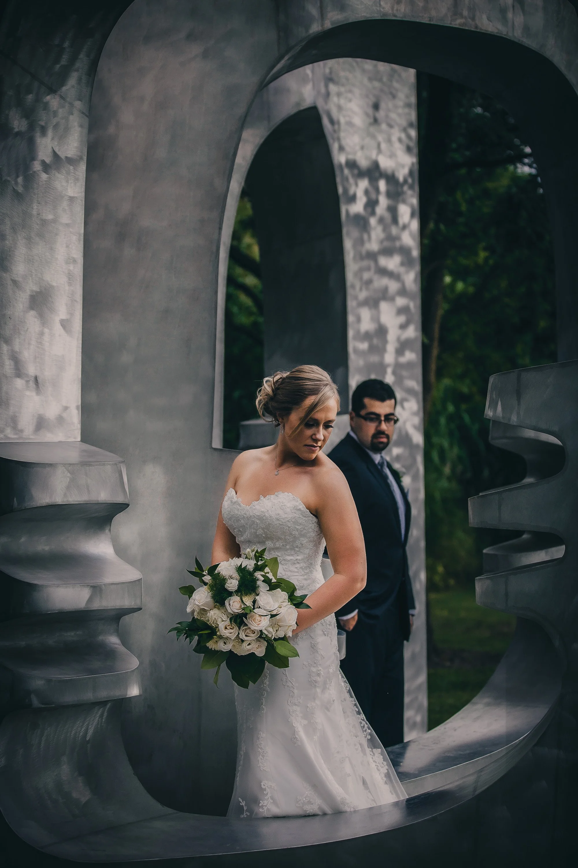 A bride in a white lace wedding gown holding a bouquet of white roses and greenery, standing outdoors next to a modern metallic sculpture with a groom in a dark suit and glasses in the background.