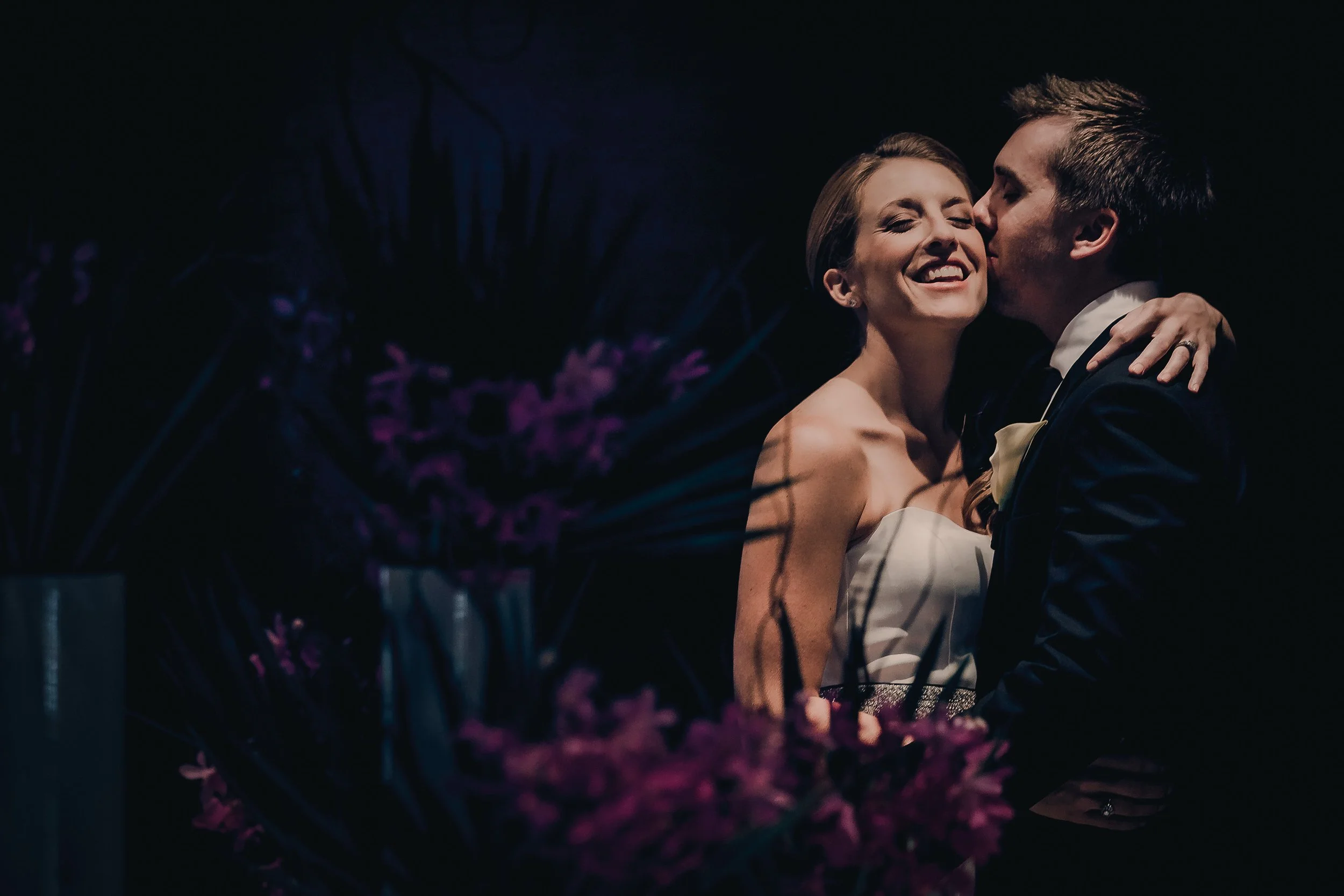 A bride and groom sharing a kiss at their wedding, with the bride smiling widely and eyes closed, wearing a white strapless wedding dress and the groom in a black suit, against a dark background with purple flowers in the foreground.