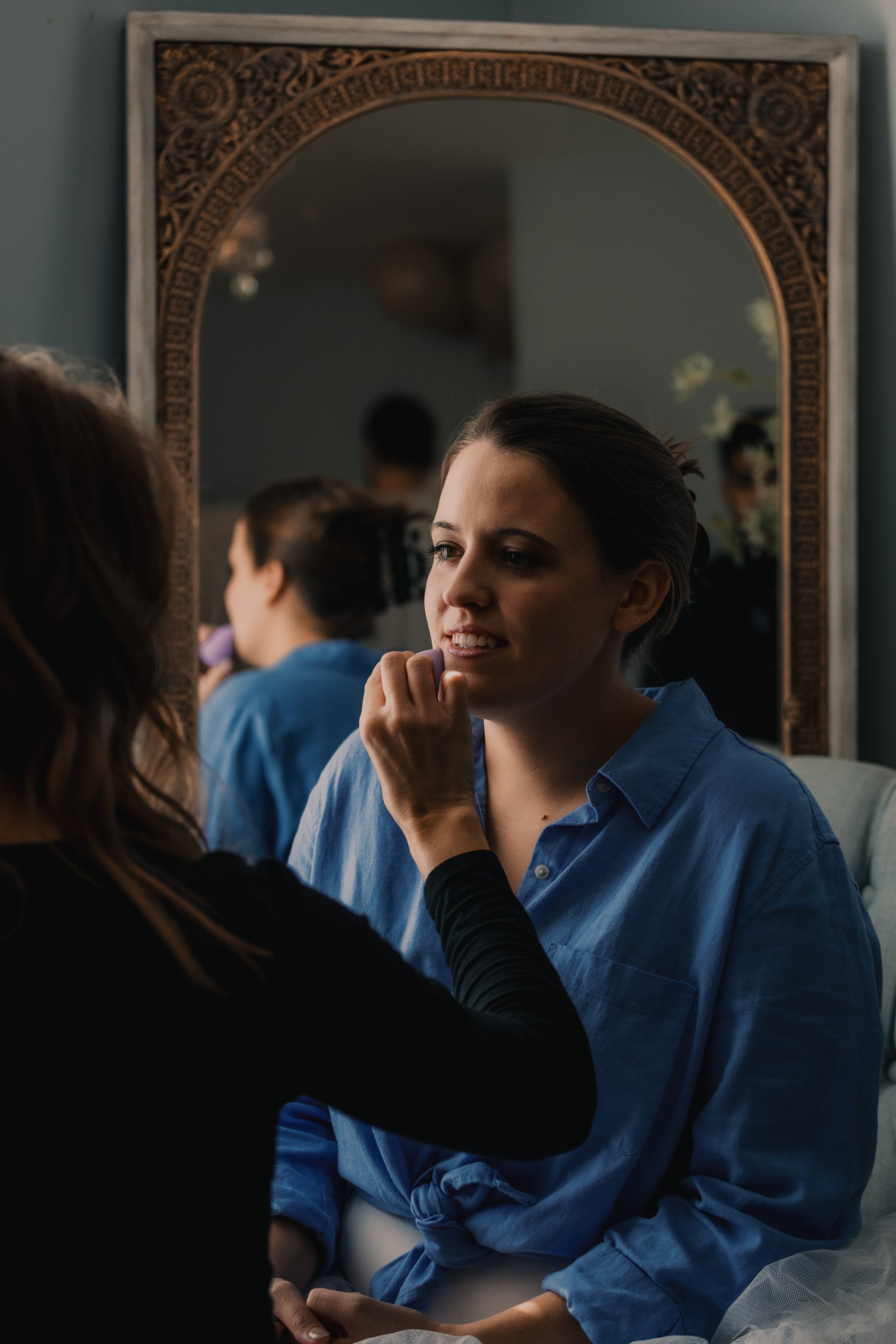 A woman in a blue shirt getting makeup applied in front of a mirror.