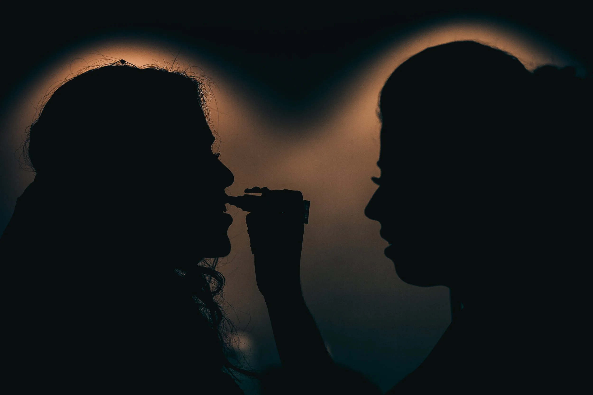 Silhouette of two people facing each other at sunset, one person holding a vape pen near the other's mouth.