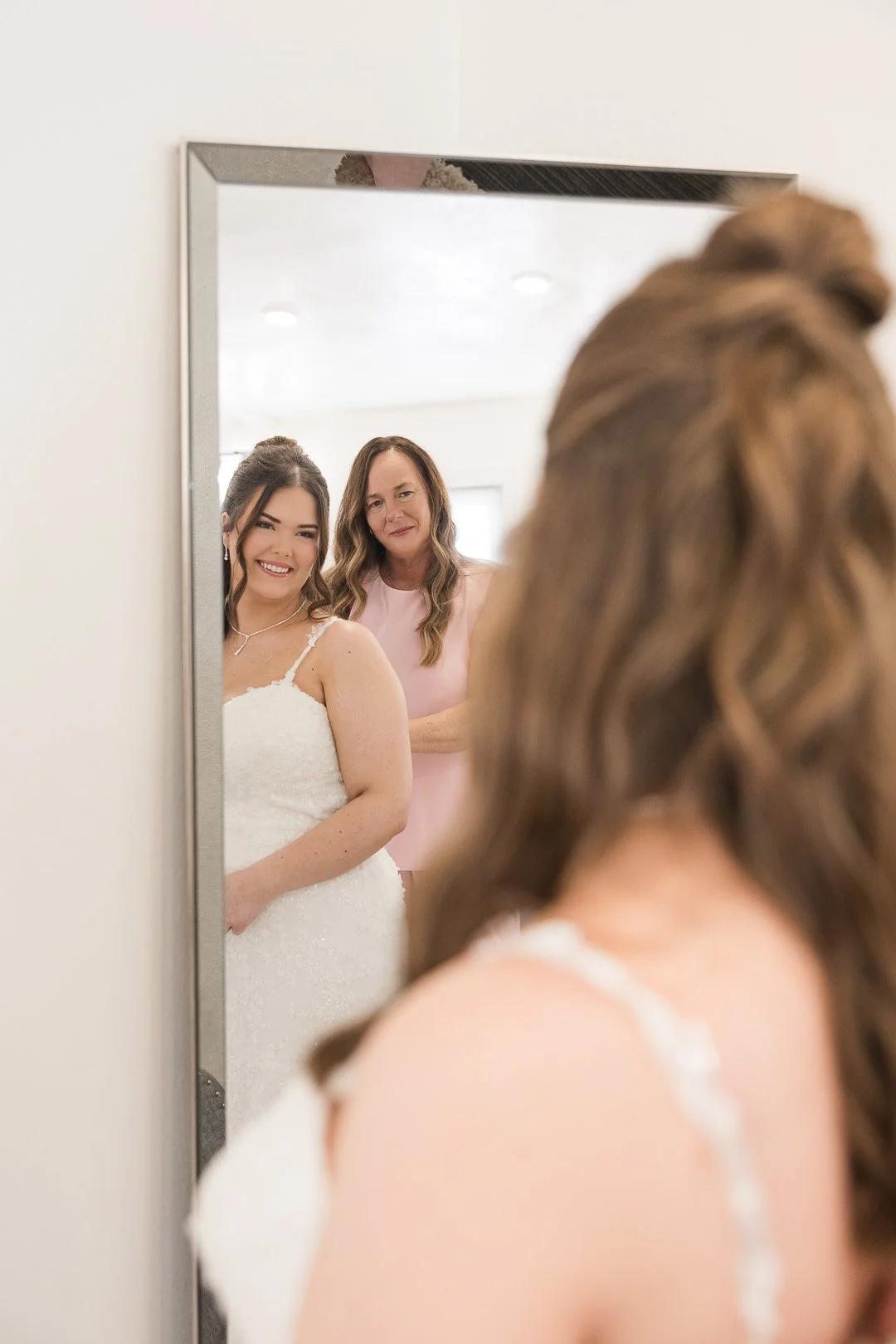 A bride in a white dress smiling as she looks into a mirror, with a woman, possibly her mother, standing behind her.