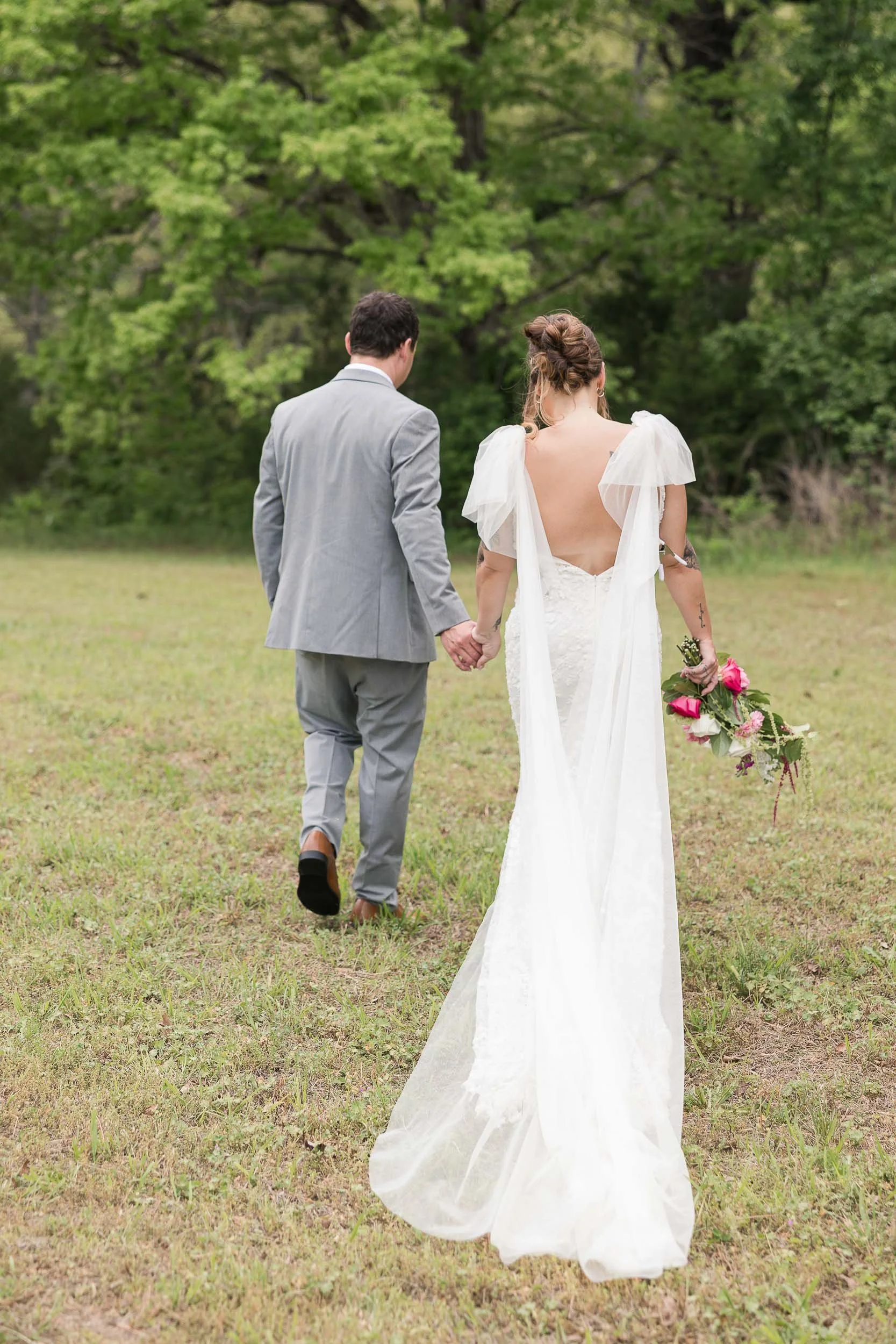Bride and groom walking on a garden path during their wedding day. The bride is holding a bouquet of flowers and wearing a white wedding dress, and the groom is dressed in a black suit with a light blue tie. They are smiling, and there are trees and greenery in the background.