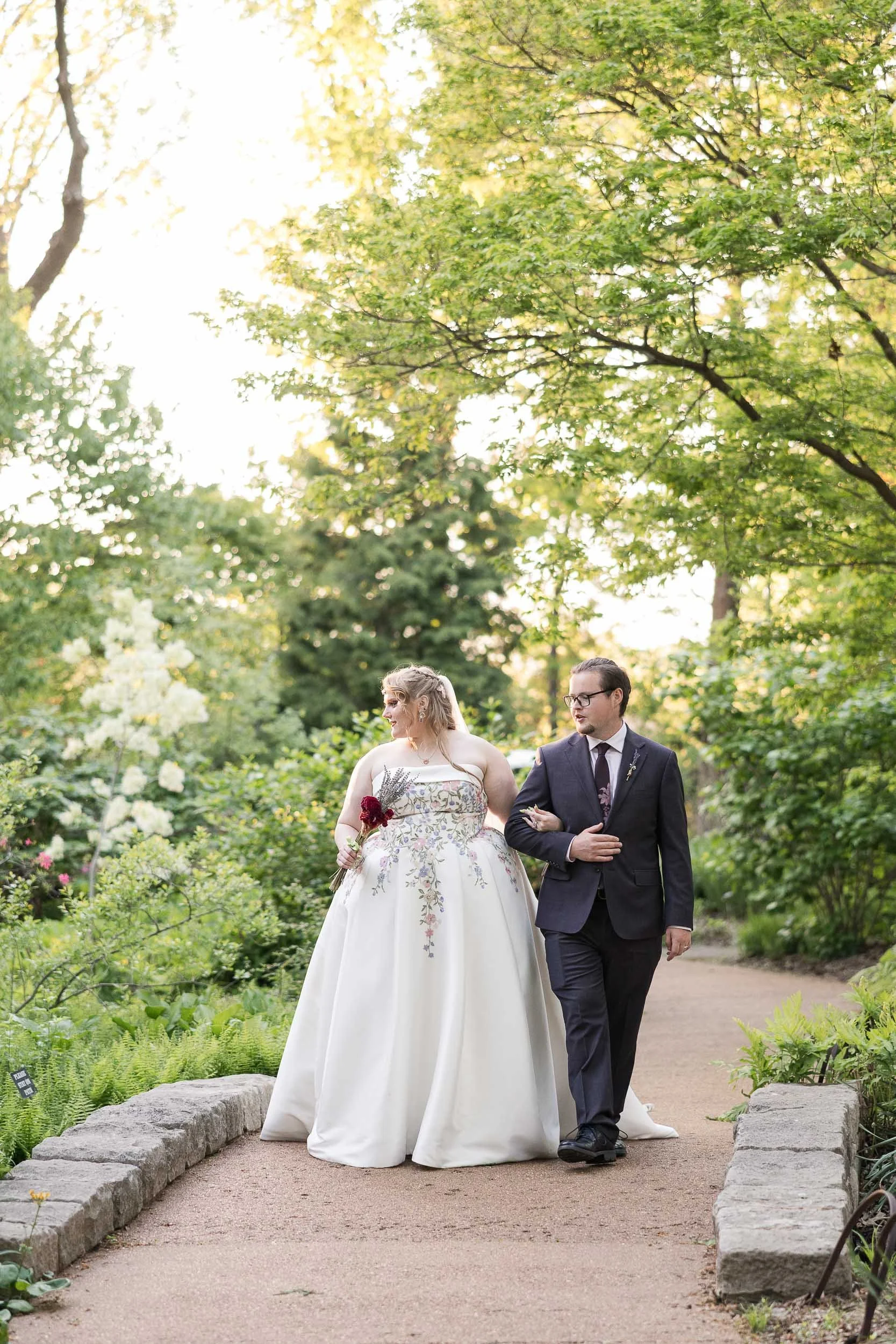 A bride and groom walking arm in arm through a green garden park during late afternoon or early evening. The bride wears a white wedding dress and holds a small bouquet, while the groom wears a dark suit and glasses.