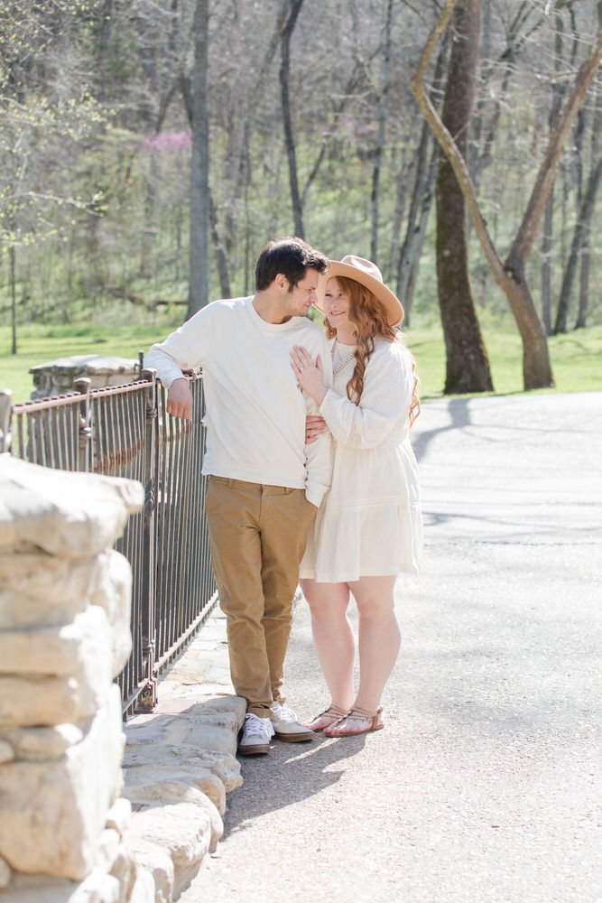 A couple stands by a railing in a park, with trees in the background. The woman is wearing a hat and light-colored dress, the man in a light sweater and pants. They look at each other affectionately.