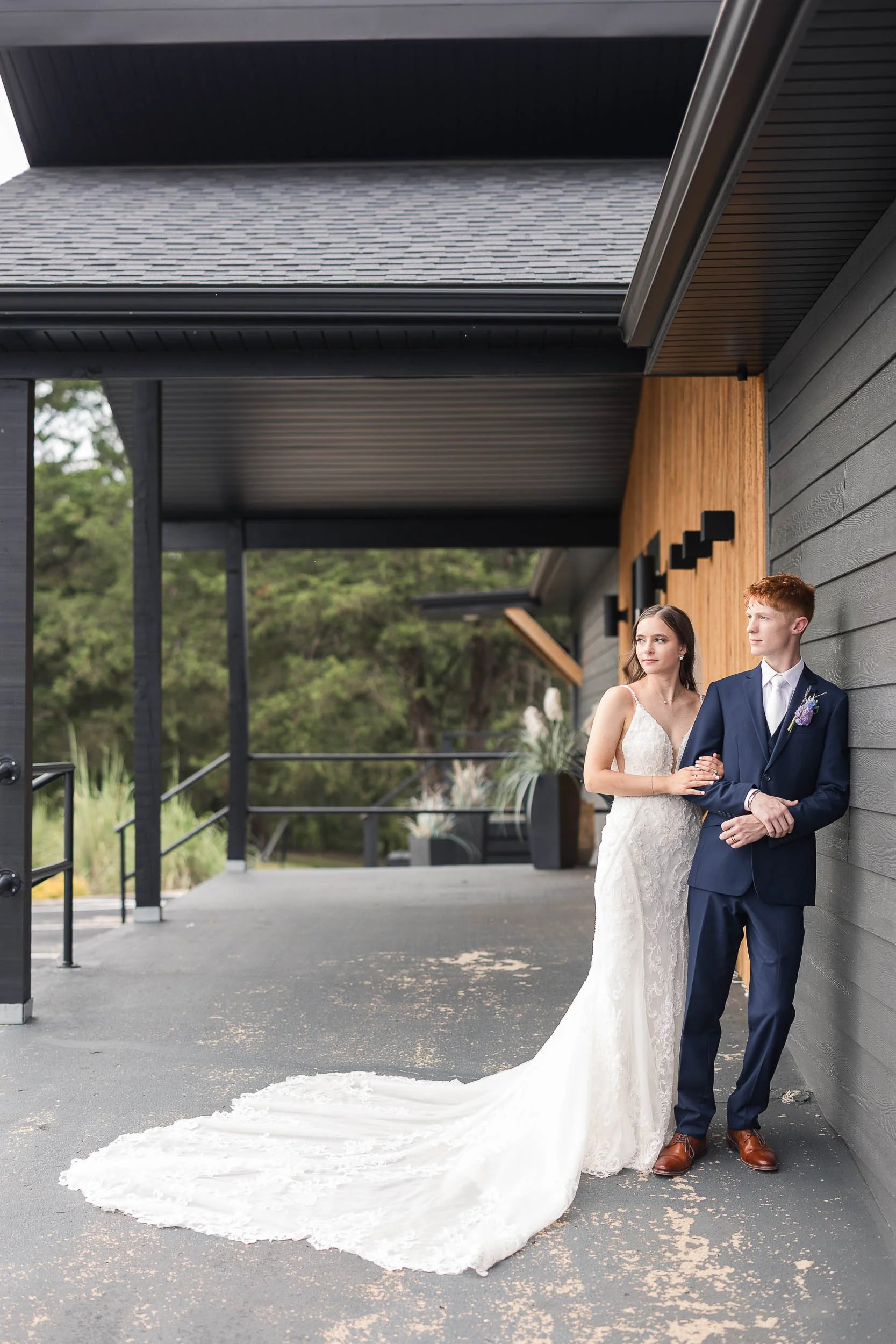 A bride and groom stand together outside a modern building, dressed in wedding attire. The bride wears a white lace wedding gown with a long train, and the groom wears a dark blue suit with a white shirt and brown shoes. They hold hands and look off 