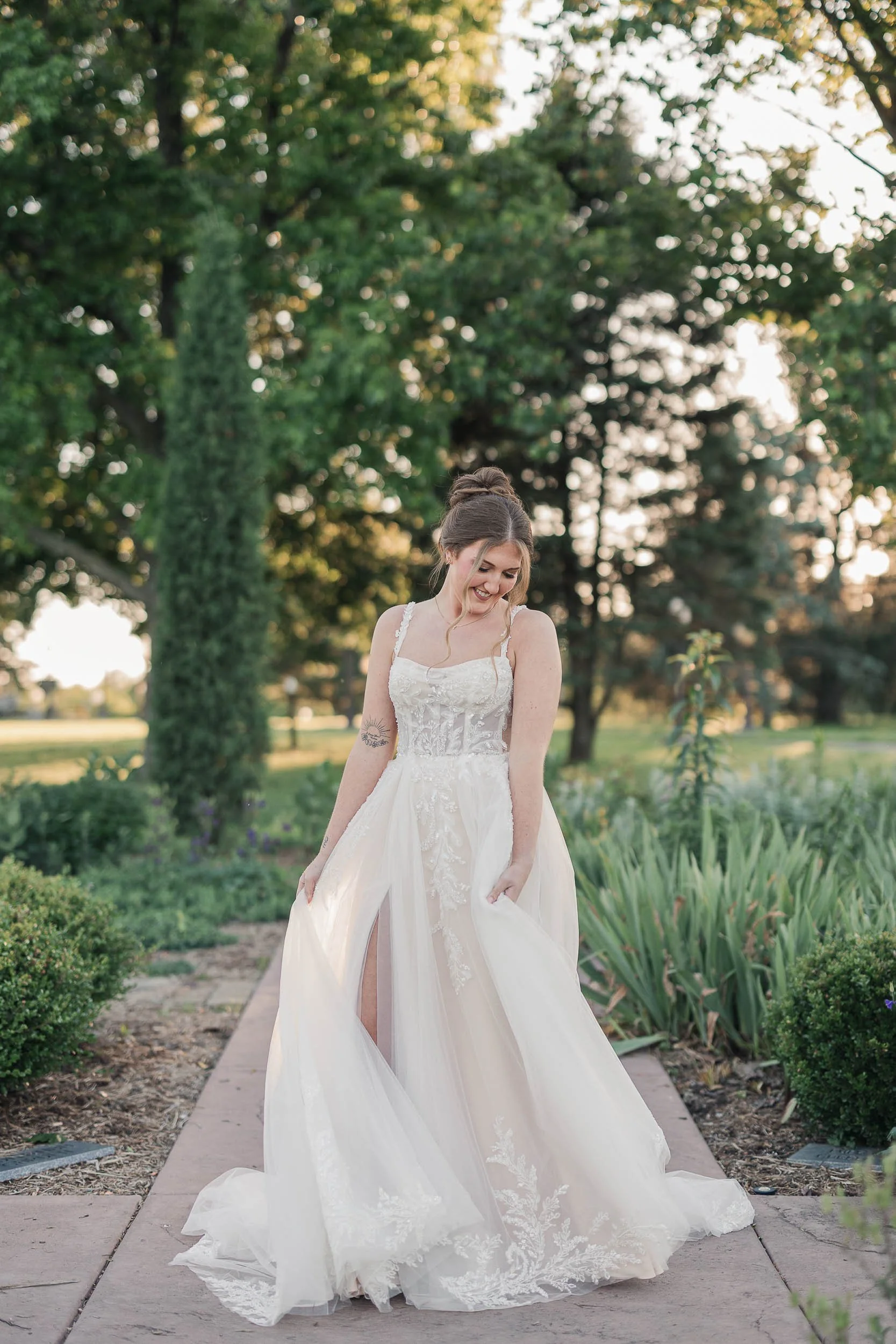 A young woman in a white wedding dress standing on a garden path, smiling and looking down, with greenery and trees in the background.