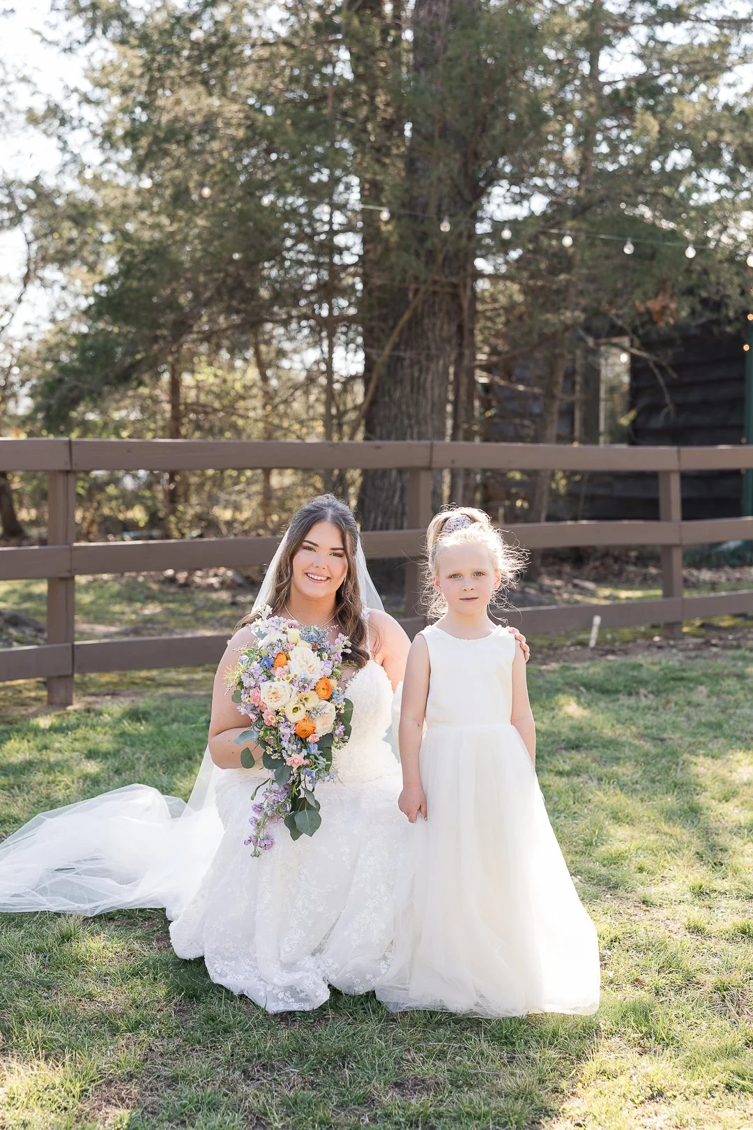 A bride in a white wedding dress kneels on grass beside a young girl in a white dress, holding a colorful bouquet of flowers, outdoors in front of a wooden fence and tall trees.