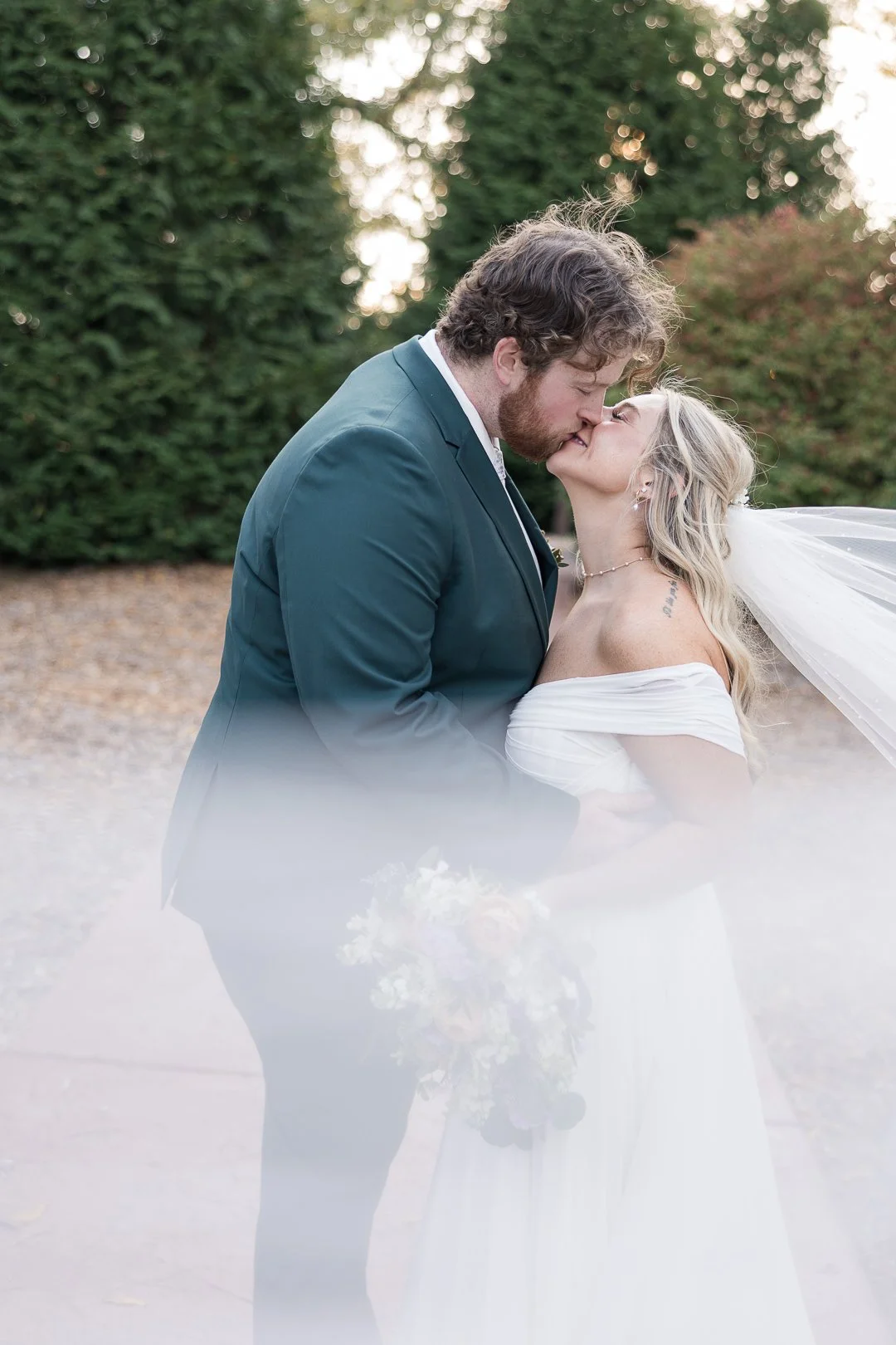 A newlywed couple sharing a kiss outdoors, the groom wearing a dark suit and the bride in a white off-shoulder wedding dress, with a bouquet in front of them and lush green trees in the background.