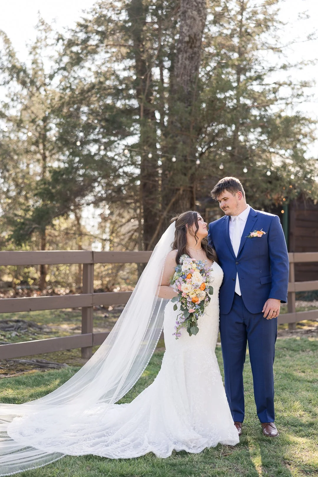 A bride and groom standing outdoors on a grassy area, with trees in the background. The bride is wearing a white lace wedding gown and veil, holding a colorful bouquet. The groom is dressed in a blue suit with a white shirt and boutonniere. They are 