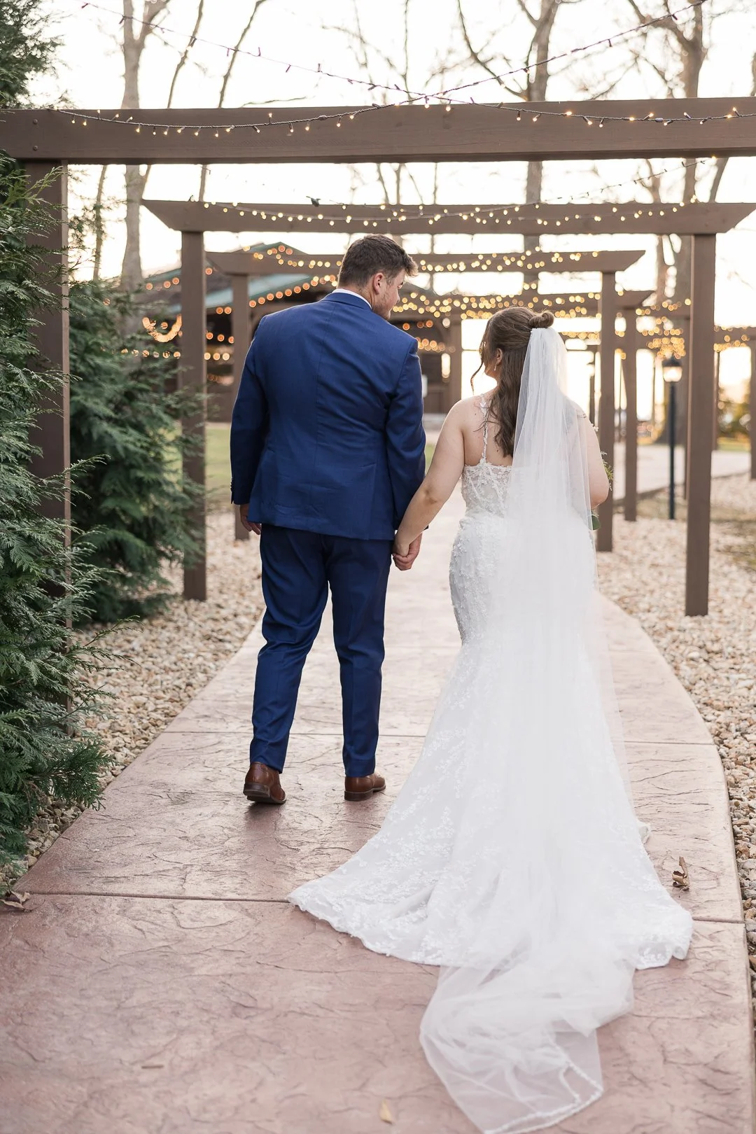 A bride and groom holding hands walking on a path decorated with string lights hanging from wooden beams, surrounded by trees and greenery, during a wedding ceremony.