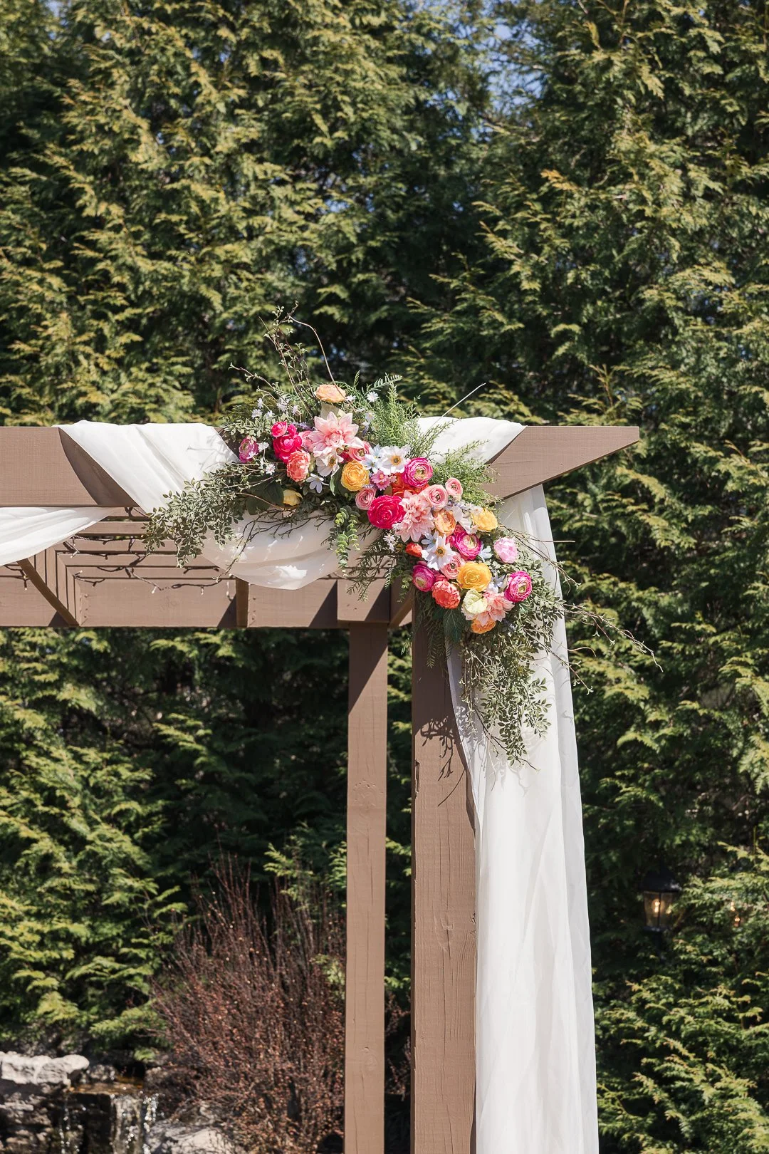 Flower arrangement with pink, yellow, and white flowers on a wooden outdoor arch decorated with white fabric, set against green trees.