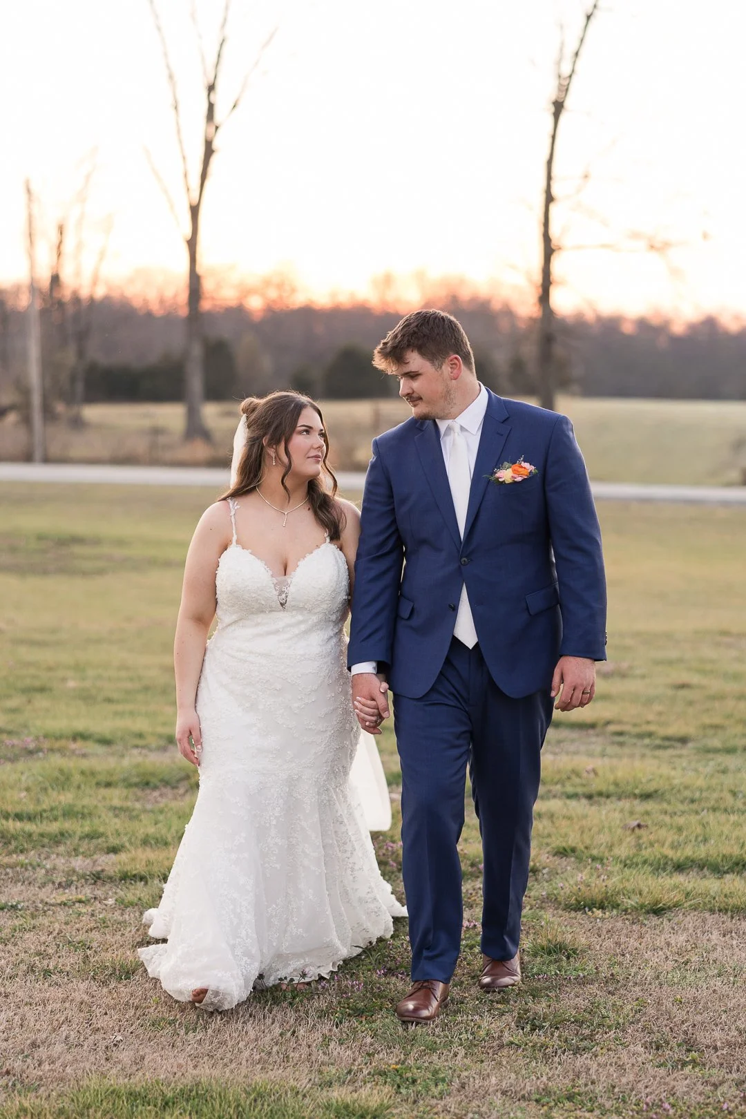 A bride and groom walking hand in hand outdoors during sunset, with empty trees in the background, dressed in wedding attire.