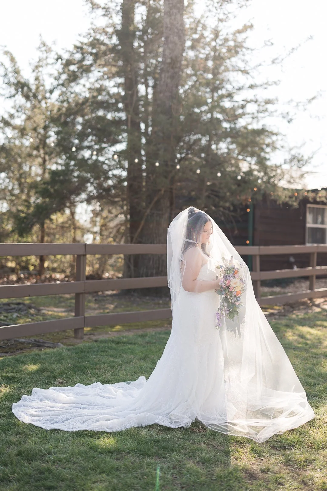 A bride in a white wedding gown with a long train and veil, holding a bouquet of flowers, standing outdoors in front of a wooden fence with trees and sunlight in the background.