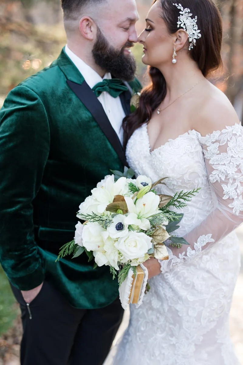 Bride and groom standing close, holding bouquet with white flowers, bride in lace gown with decorative headpiece, groom in green velvet suit.