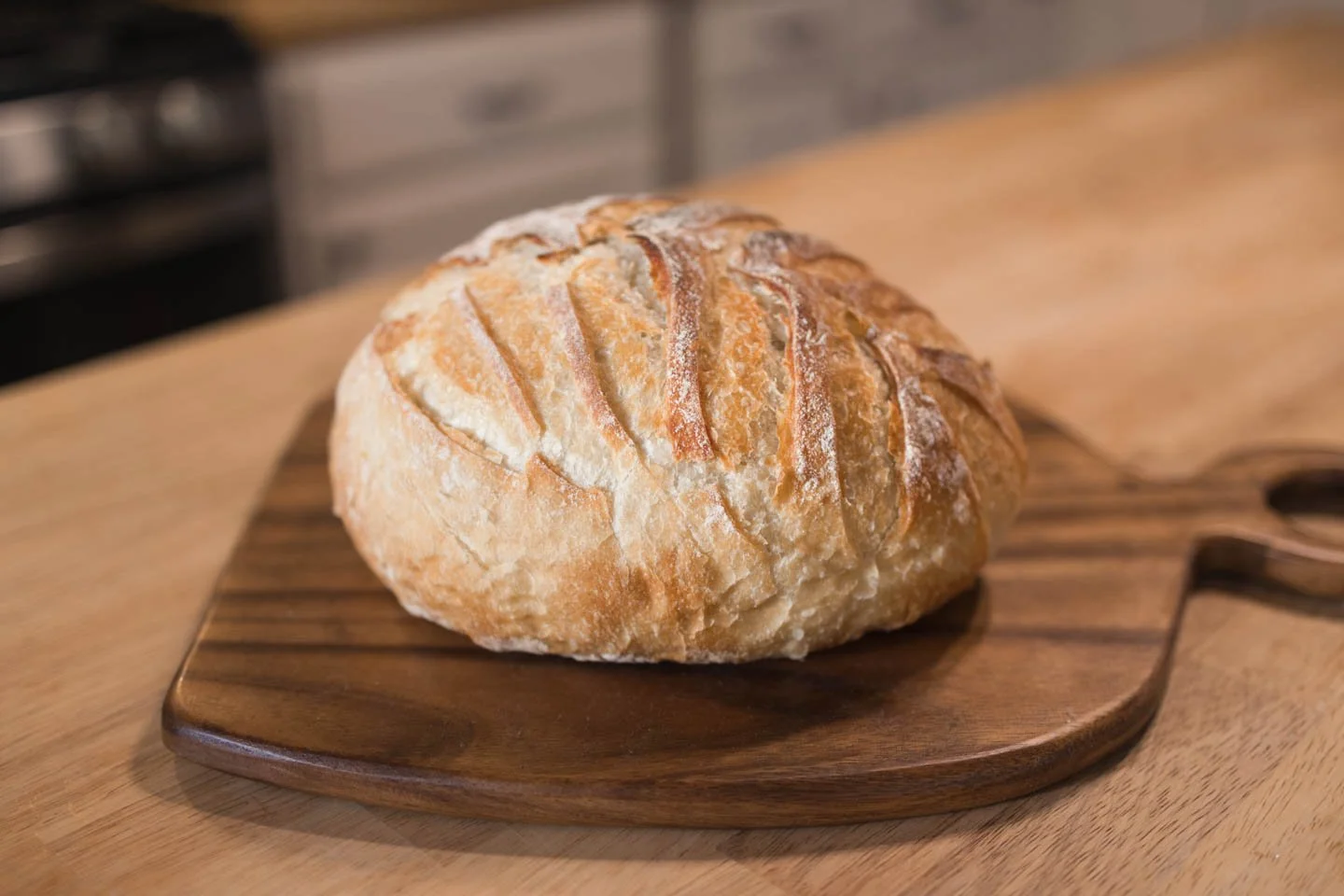A round loaf of bread with a rustic, crusty exterior, scored on top, sitting on a wooden cutting board.