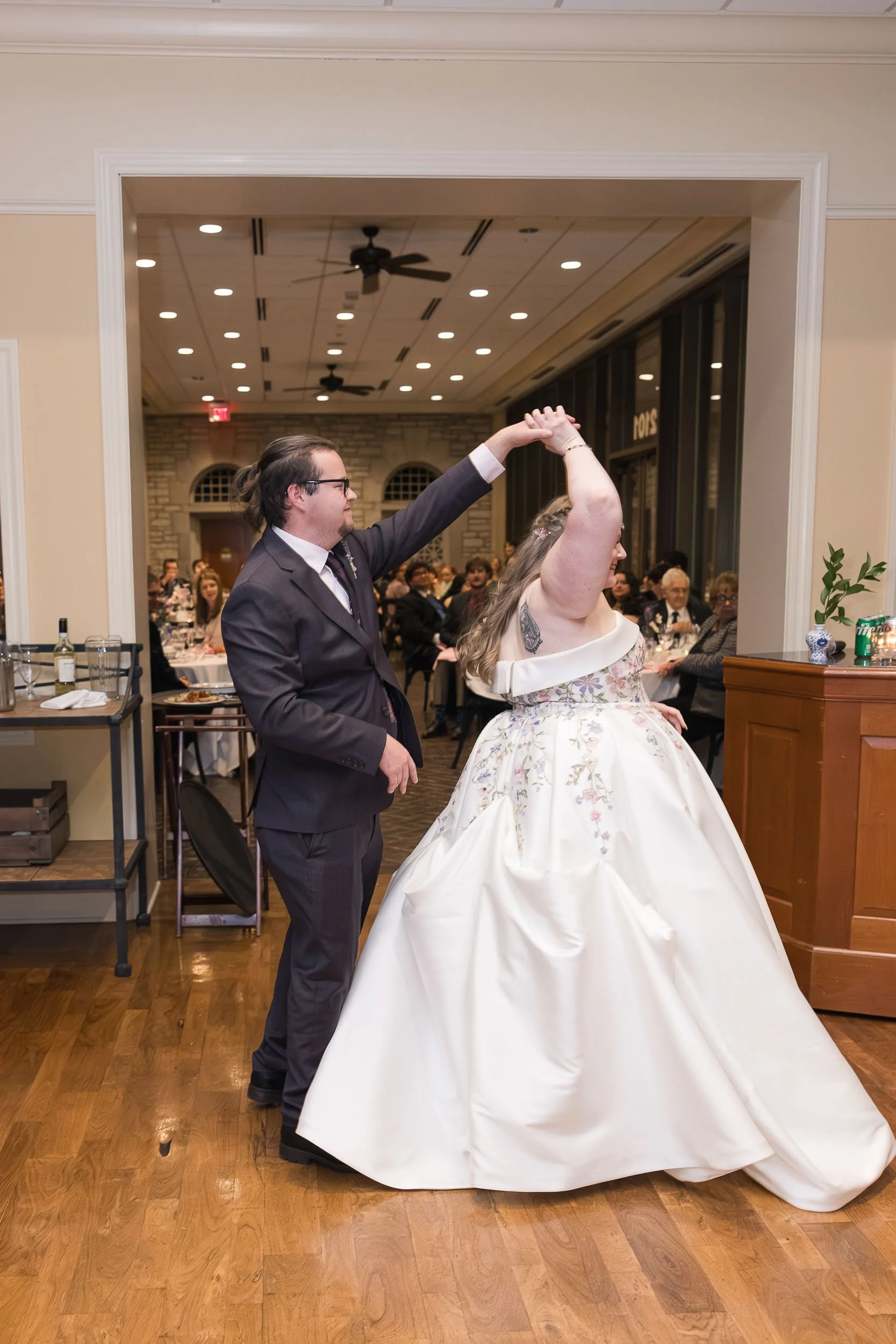 A bride and groom dancing during their wedding reception. The groom is wearing a black suit and glasses, and the bride is in a white floral wedding gown. They are holding hands and dancing in a decorated banquet hall filled with seated guests.
