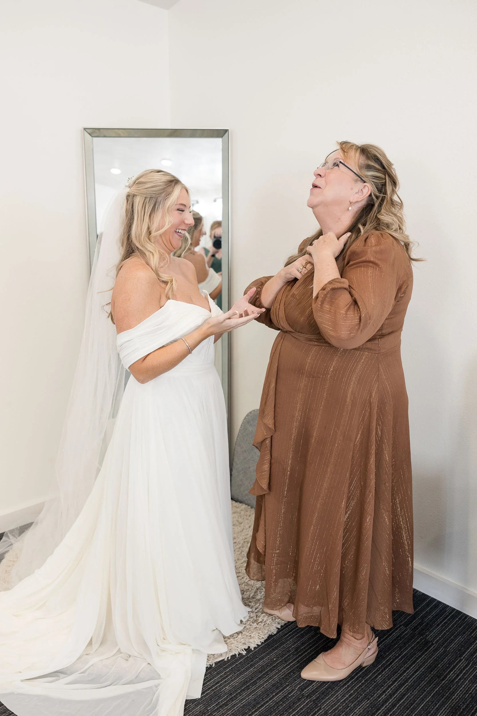 A bride in a white dress and veil smiling and laughing while talking with a woman in a brown dress and glasses. They are in a room with a mirror, cream-colored walls, and a black and gray carpet.