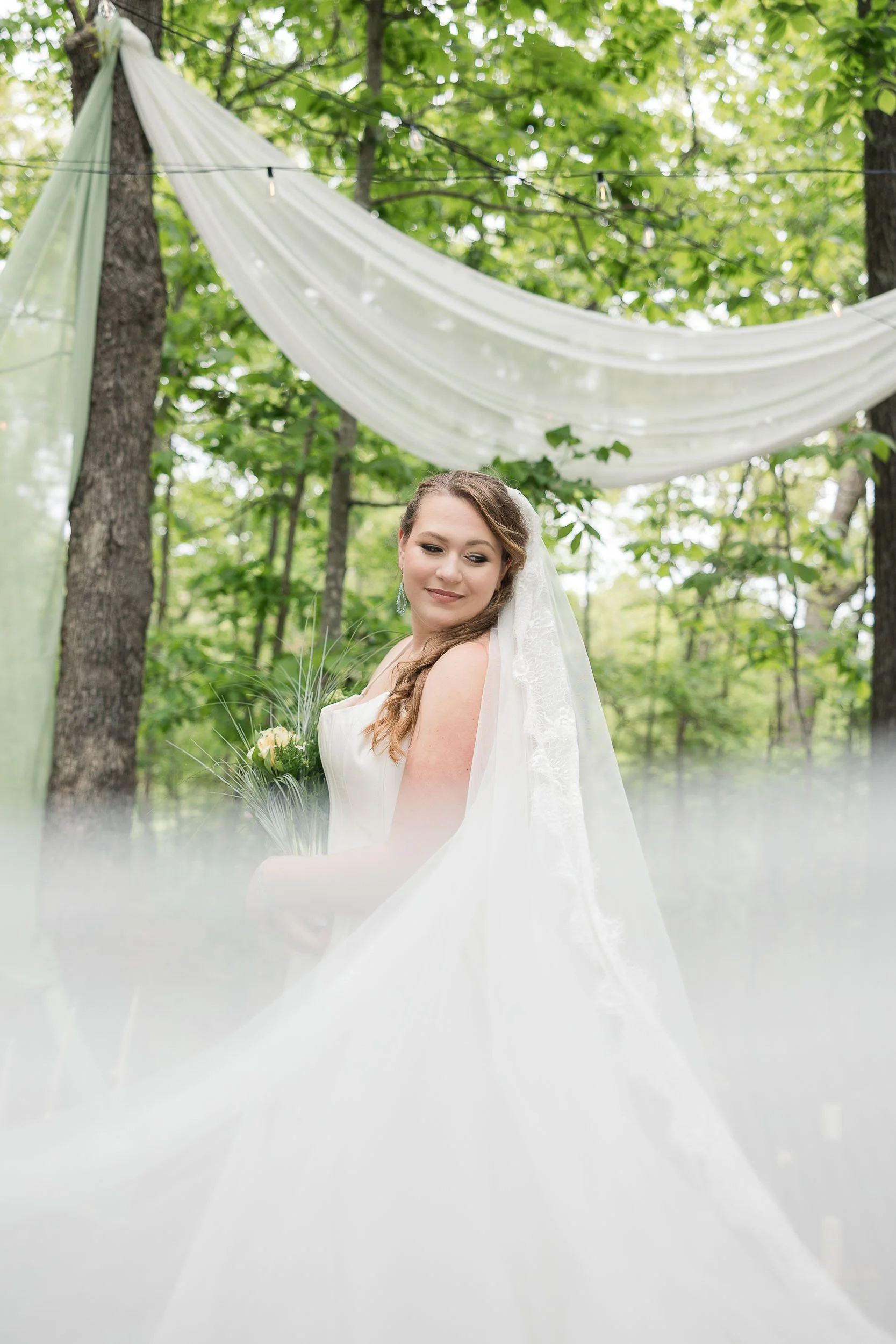 A bride in a white wedding dress holding a bouquet, standing outdoors in a wooded area with green leaves and trees, under a draped white fabric and string lights.