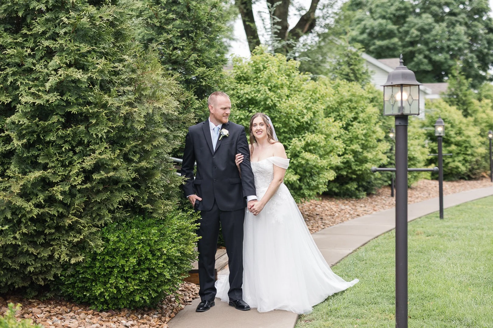 A bride and groom holding hands and smiling outdoors on their wedding day, standing on a sidewalk with trees and lampposts in the background.