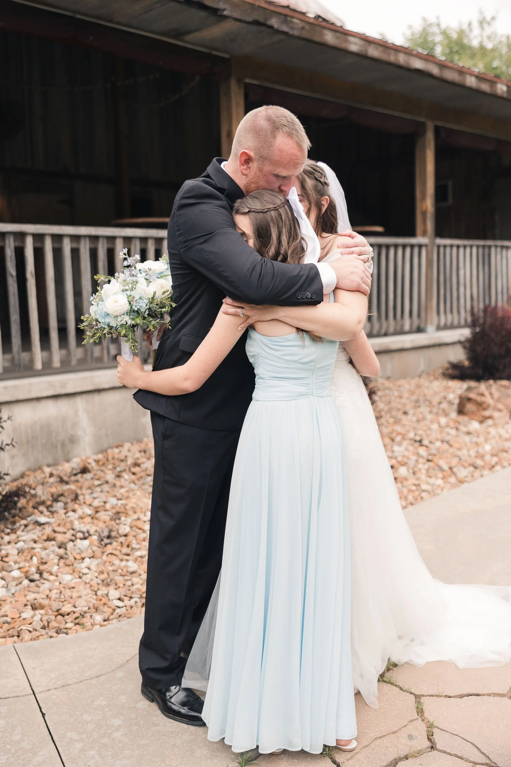 A groom in a black suit and bride in a light blue dress embrace each other during a wedding outdoor ceremony, with a woman in a white veil also hugging the bride. The setting features a rustic wooden building and a pathway lined with rocks.