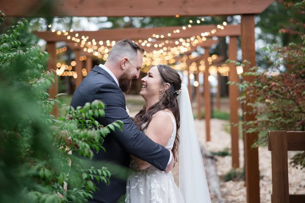 Bride and groom smiling at each other under wooden pergola with string lights during outdoor wedding ceremony.